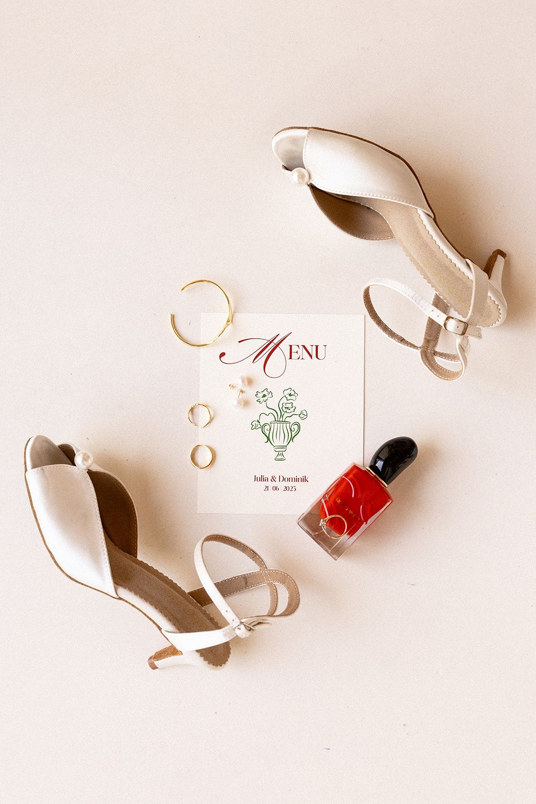 Wedding details flatlay with white heels, gold rings, red nail polish, and a delicate menu card with floral design.