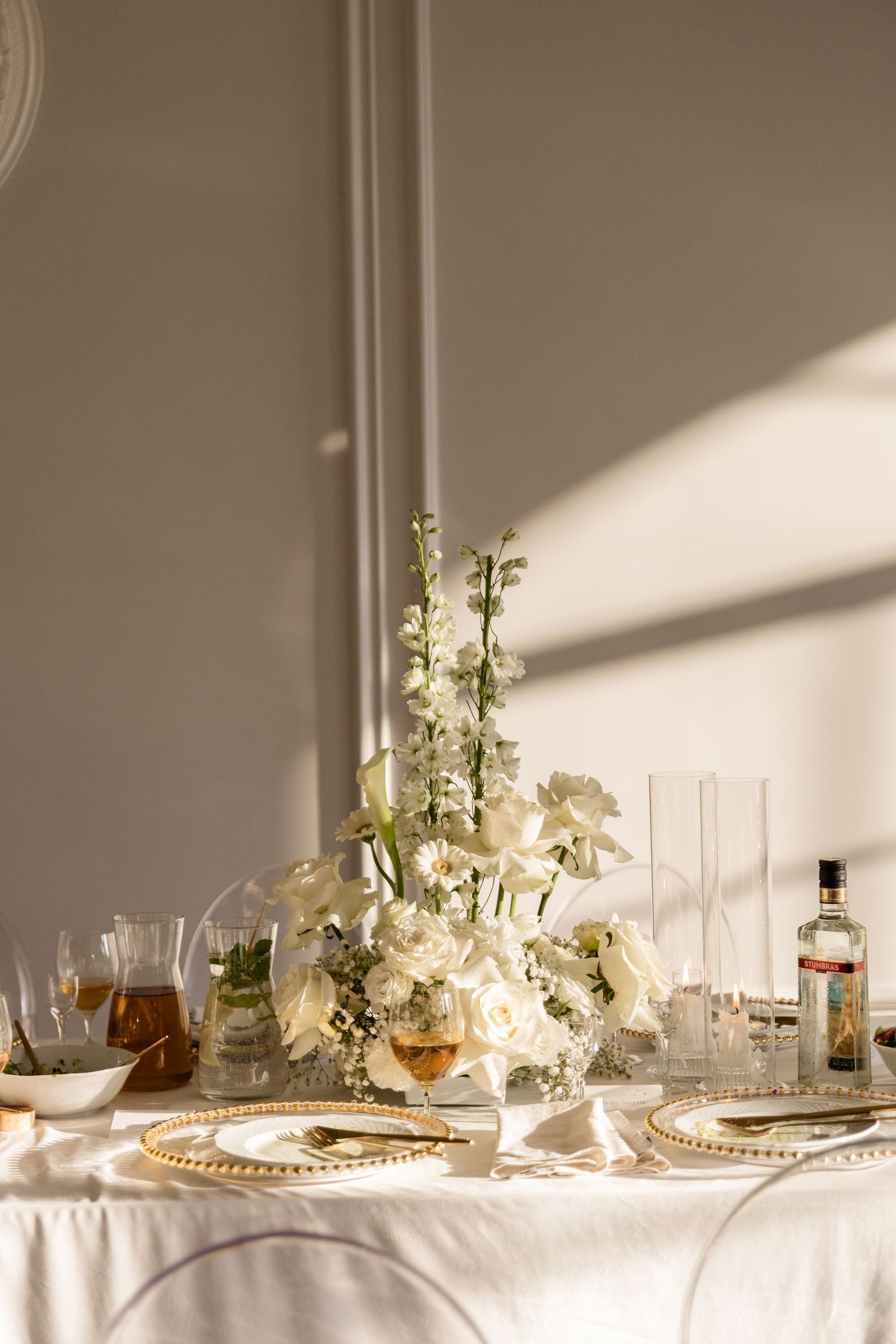 Elegant table setting with white flowers, crystal glassware and golden charger plates in soft natural sunlight.