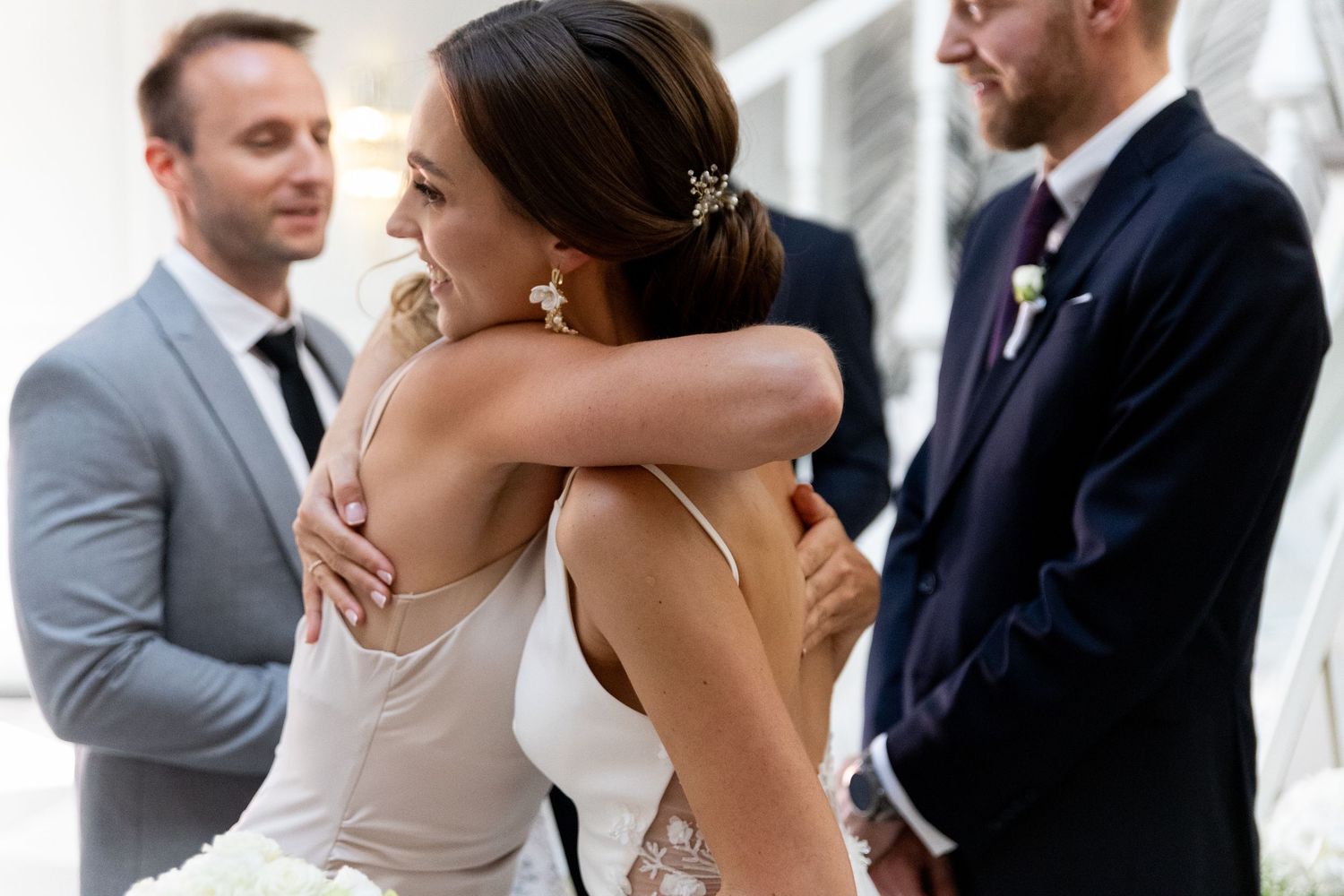 Two guests embrace warmly at a formal wedding celebration as others look on in elegant attire.