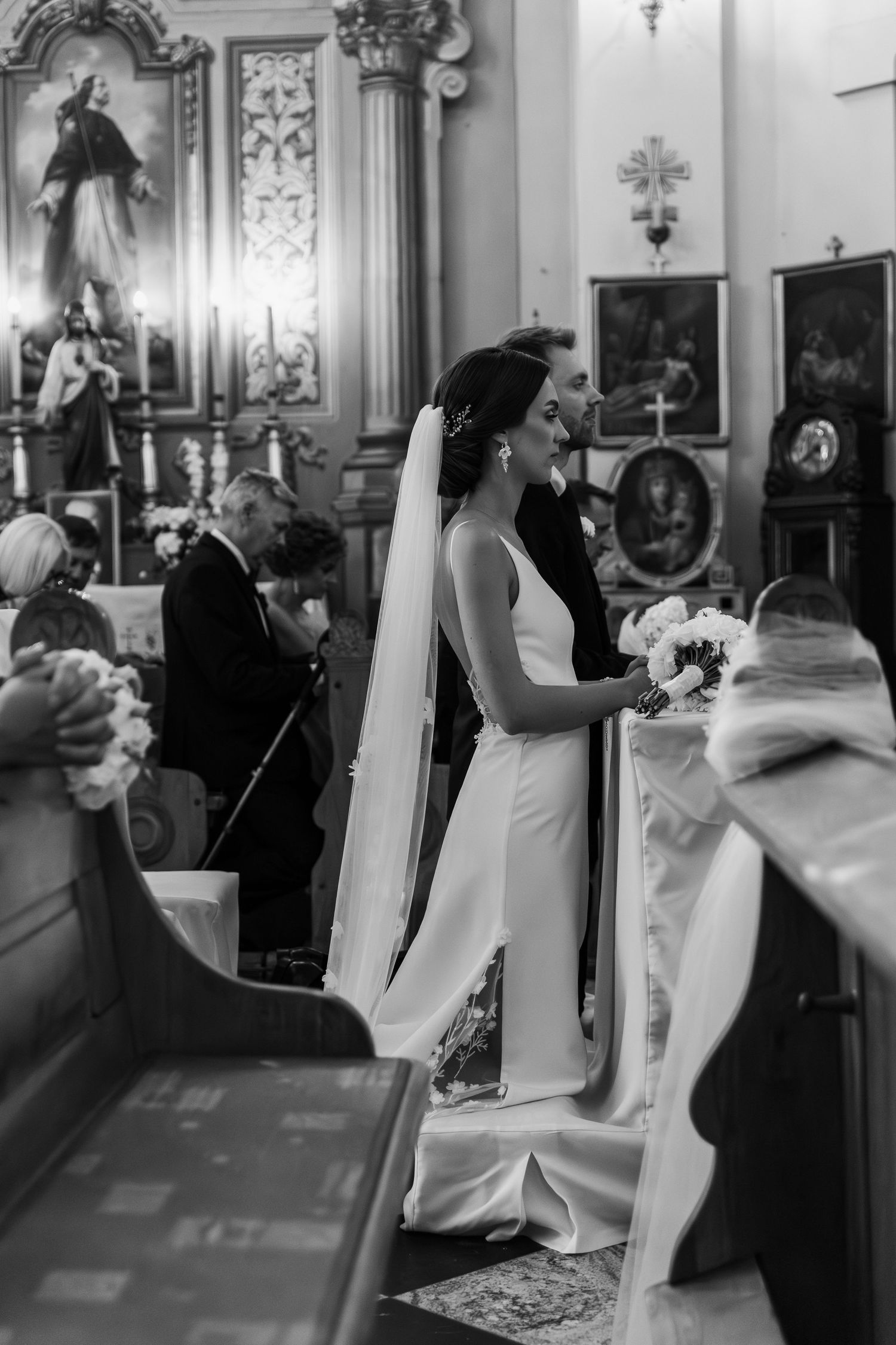 A black and white wedding ceremony photo showing people inside an ornate church with elegant decor and religious artwork.