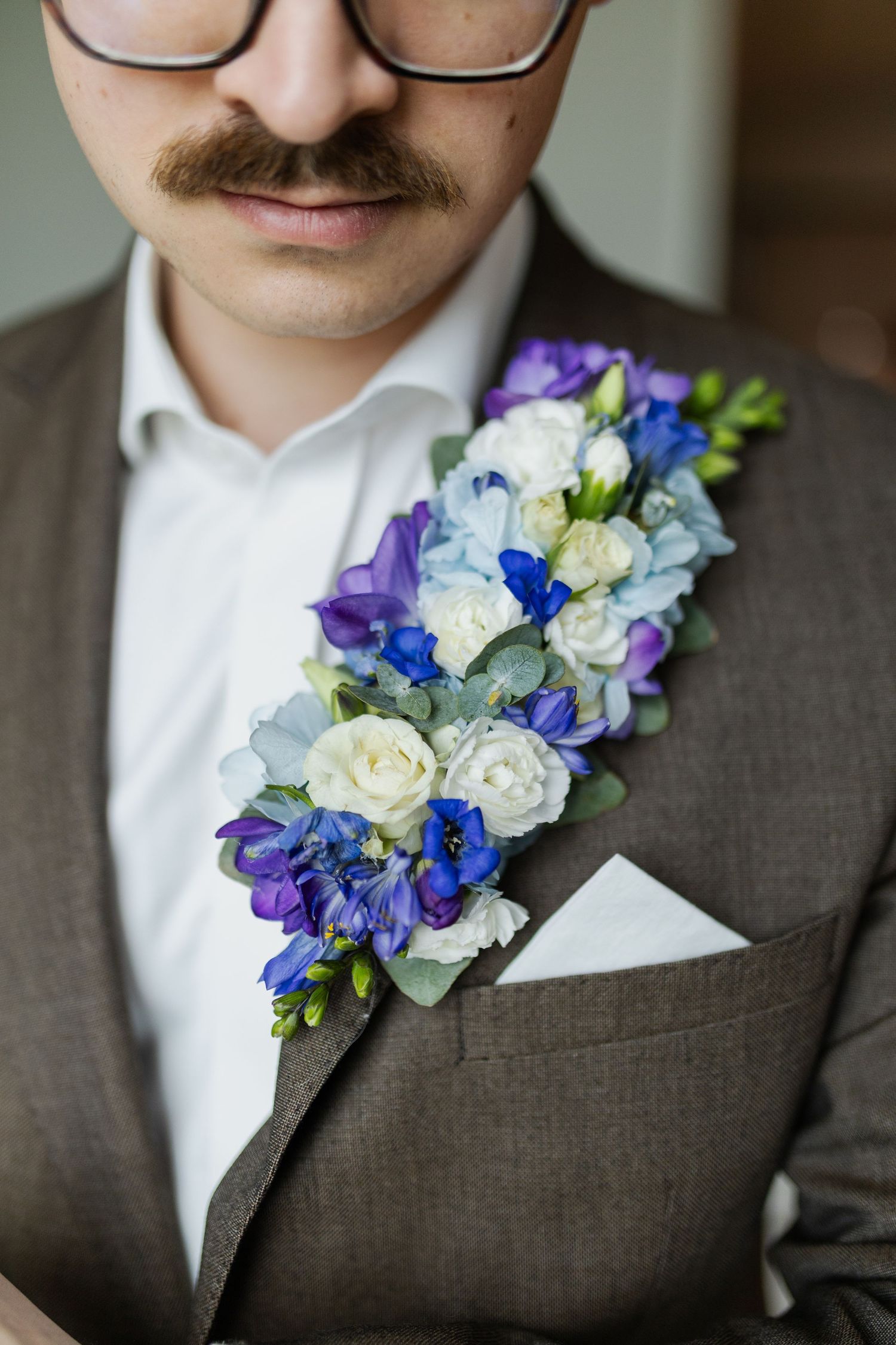 A wedding boutonniere with purple, blue and white flowers adorns a brown suit jacket with pocket square.