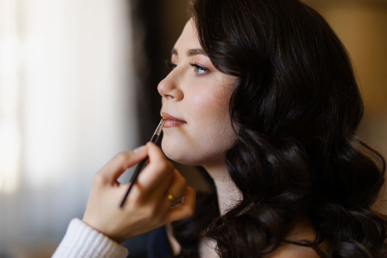 A side profile view shows black wavy hair while someone applies makeup with a brush in soft lighting.