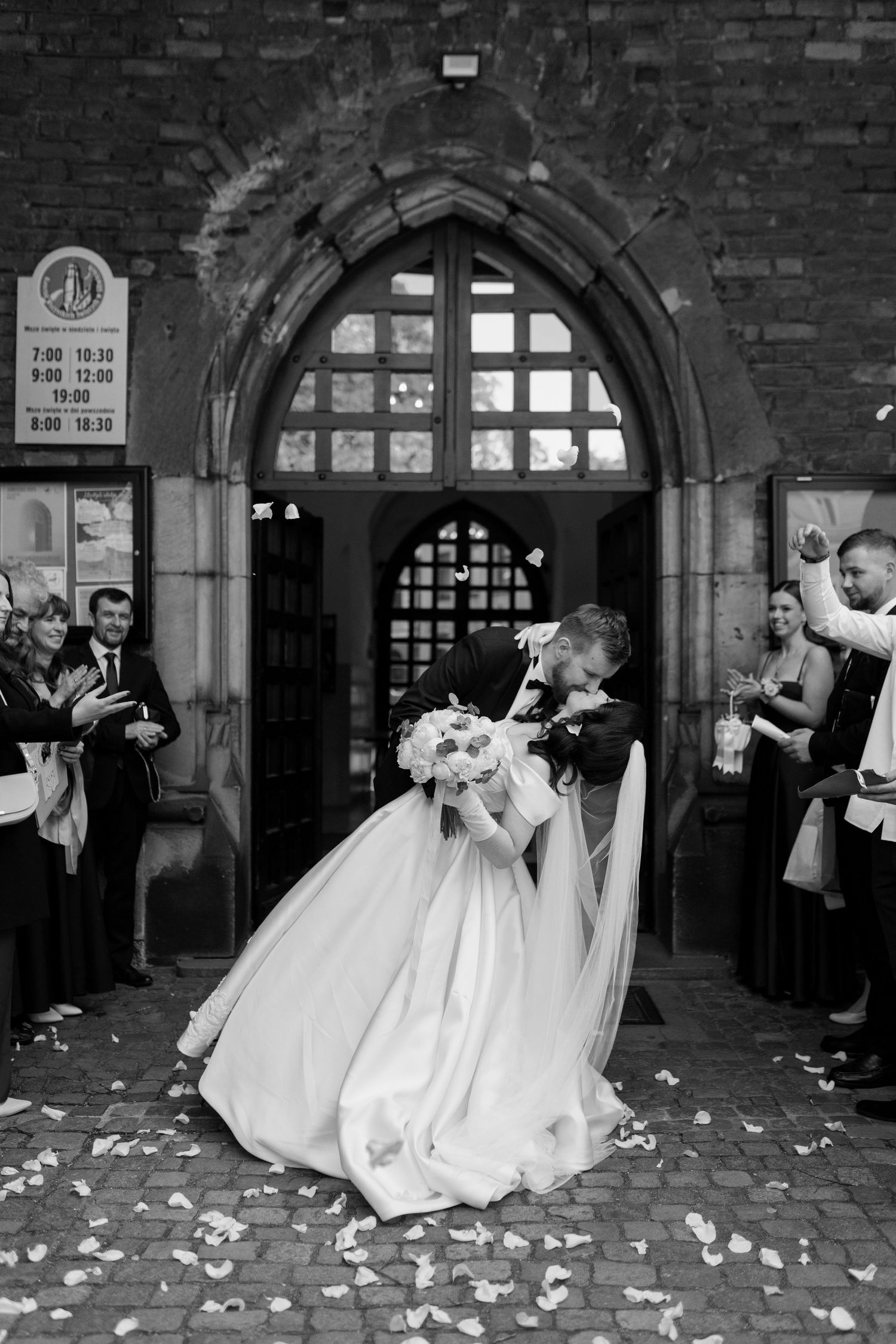 A romantic black and white wedding photo showing the newlyweds embracing in front of an arched church doorway with flower petals.
