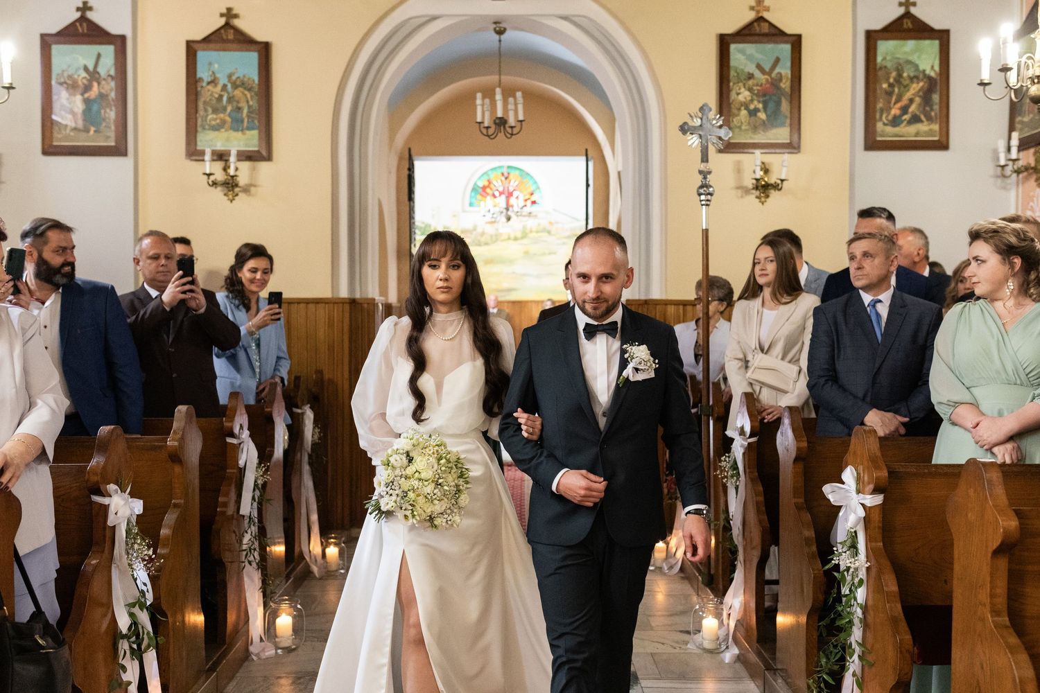 A couple walks down the church aisle after their wedding ceremony as guests look on from wooden pews decorated with flowers.