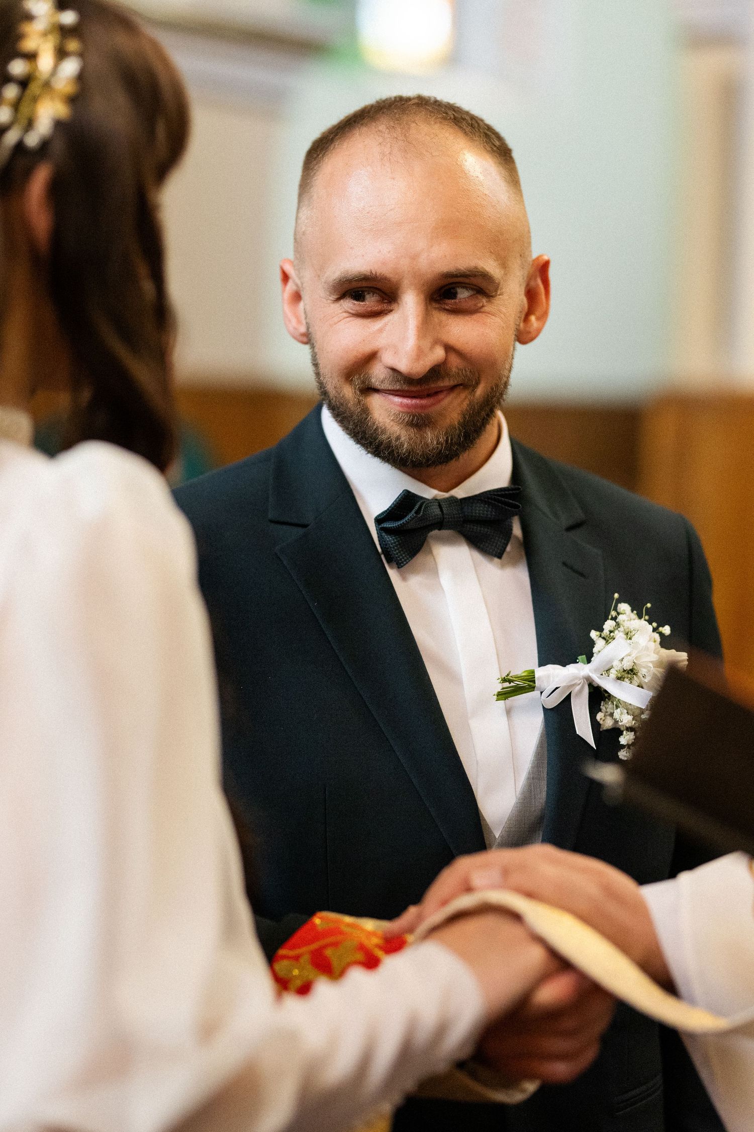Wedding ceremony in a church with a groom wearing a tuxedo and bow tie smiling warmly during the exchange of vows.