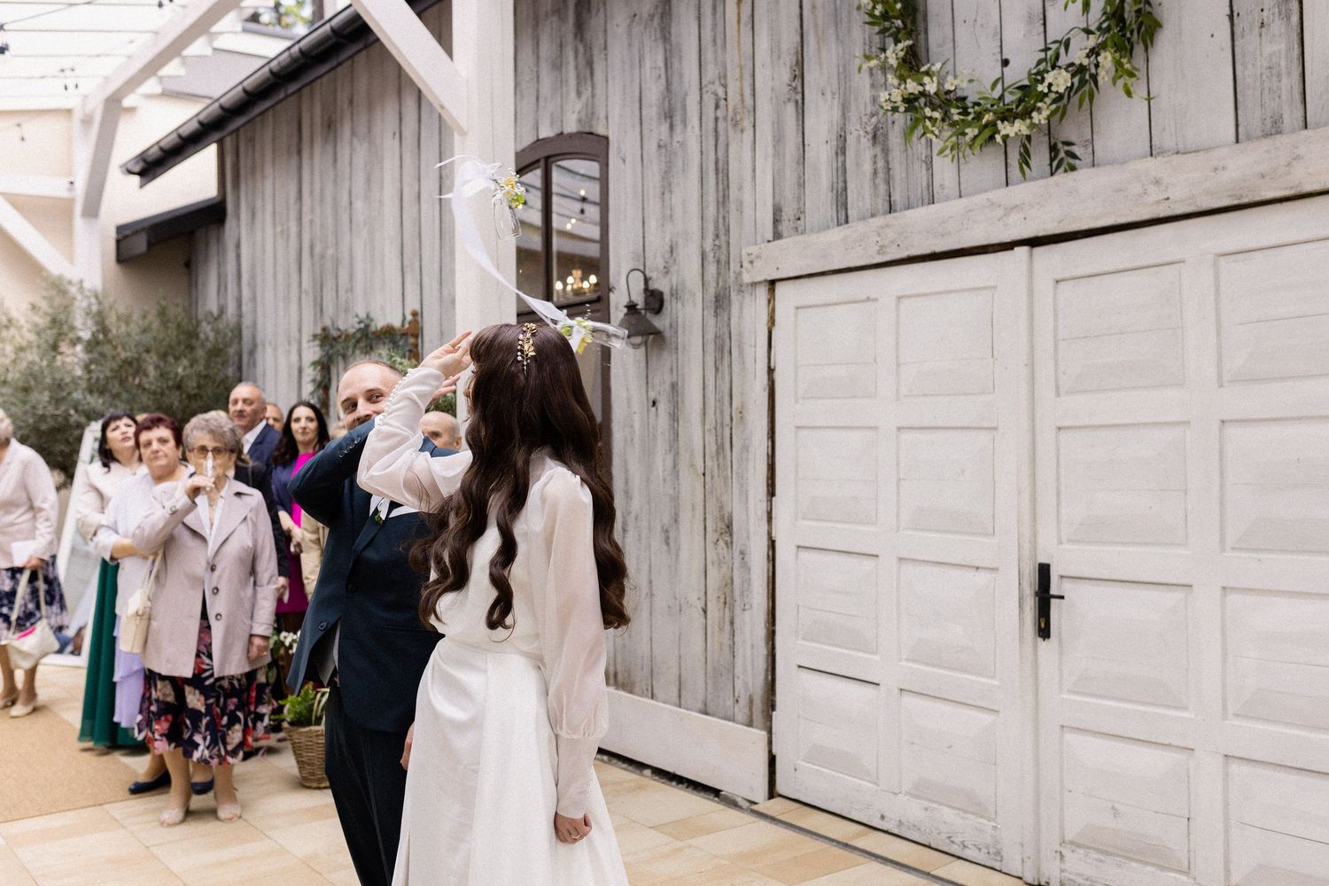A couple shares a romantic kiss in front of a rustic barn door with wedding guests looking on.