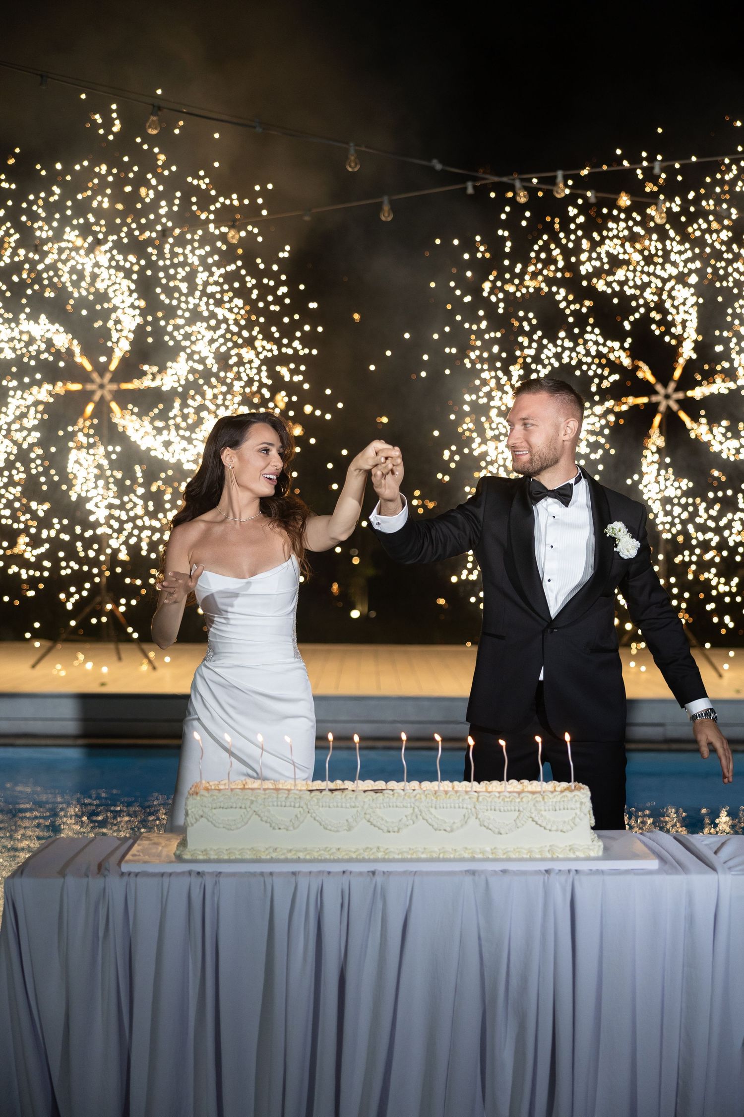 Bride and groom cutting wedding cake with sparklers glowing in background at elegant evening reception.