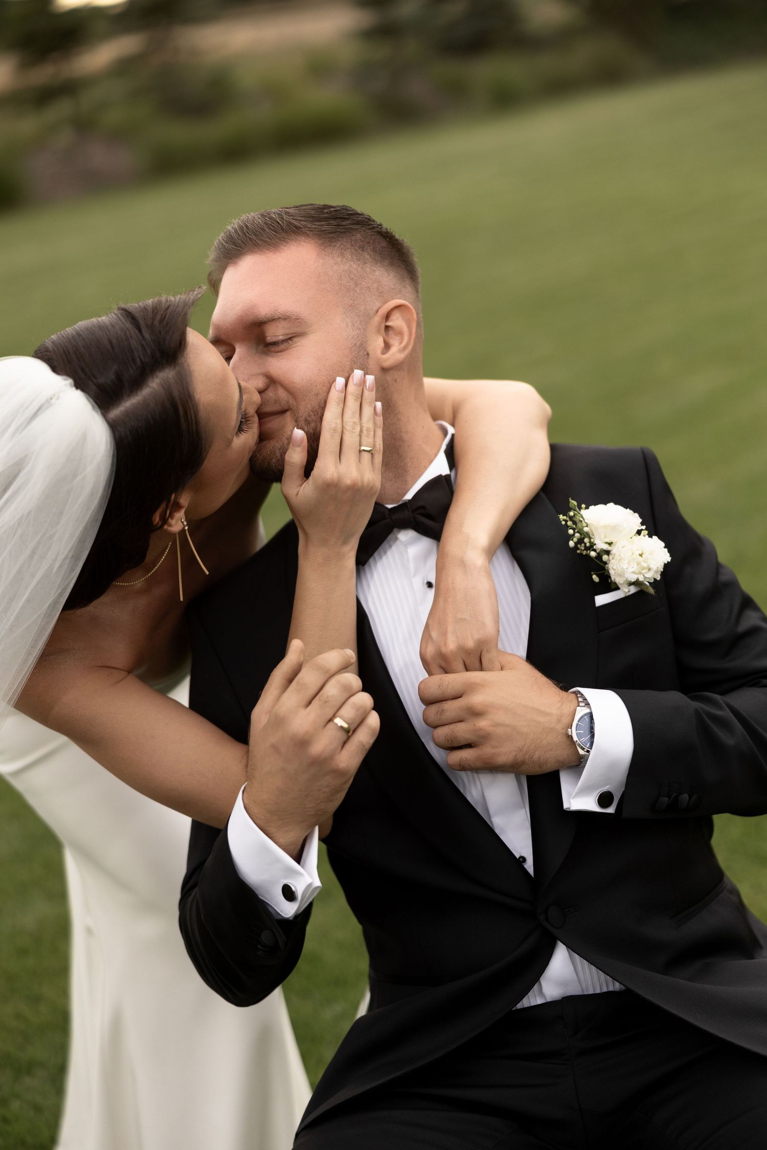 Bride and groom sharing a romantic kiss outdoors, bride showing wedding ring, groom in black tuxedo with white boutonniere.
