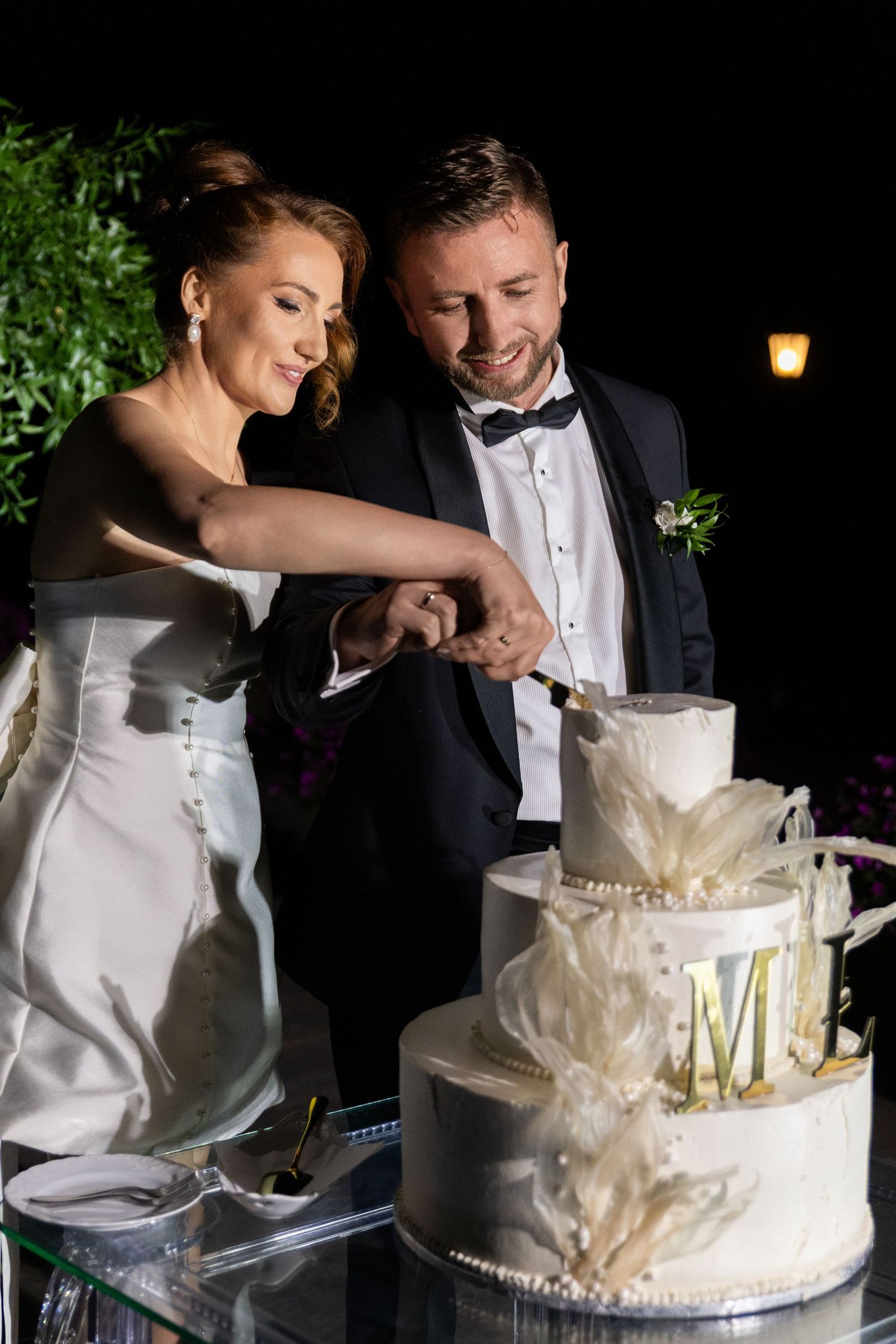 A couple shares a playful moment while cutting their elegant white wedding cake decorated with floral details.