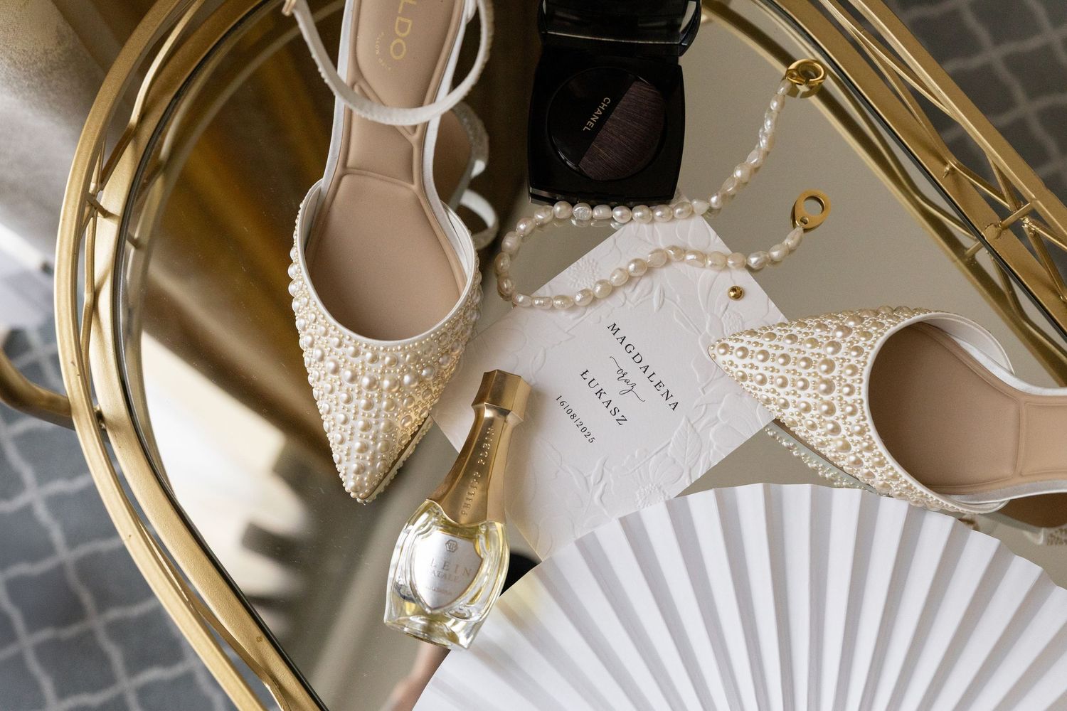 Elegant bridal accessories including pearl-adorned shoes, a delicate hand fan, and golden jewelry arranged on a mirrored surface.