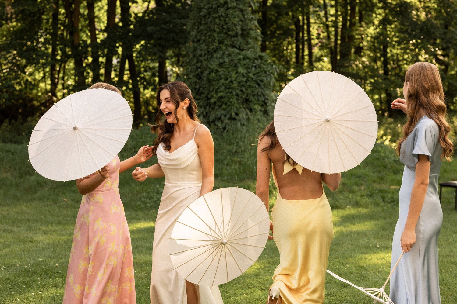 A group of bridesmaids in pastel dresses pose together with white paper parasols in a sunlit garden setting.
