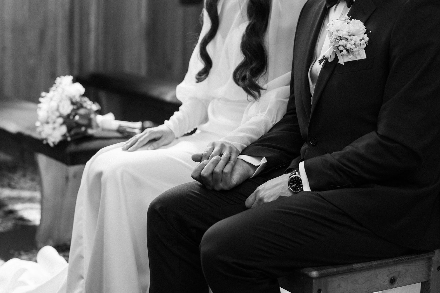 A black and white wedding photo showing a couple sitting together holding hands, with bouquet and boutonniere visible.