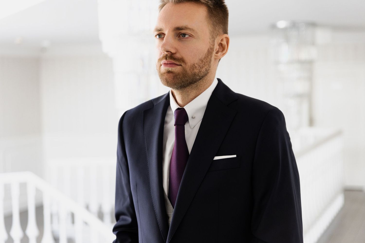 A person wearing a well-fitted black suit with white dress shirt and purple tie stands in an elegant white interior.