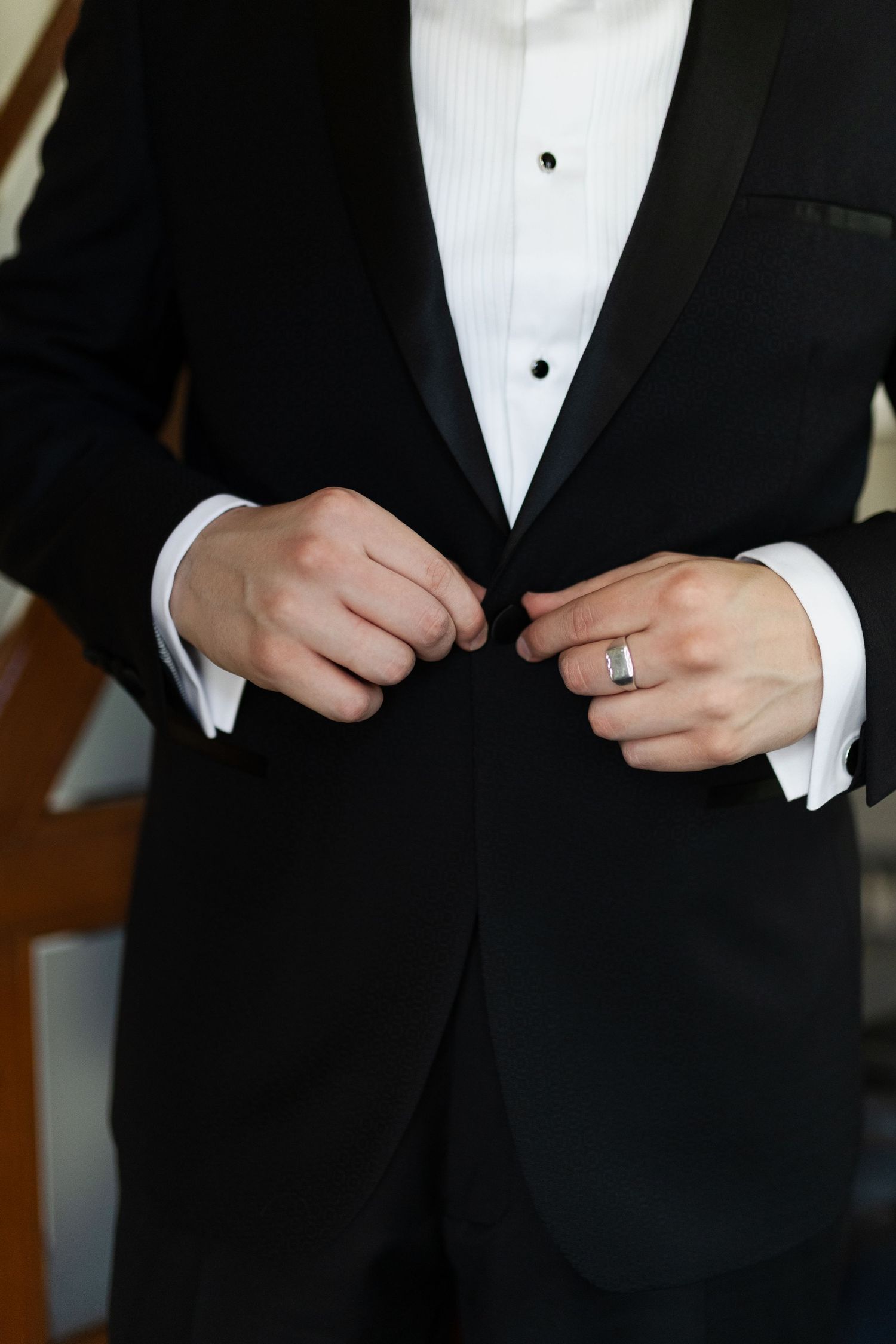Close-up of hands buttoning a formal black tuxedo jacket over white dress shirt with bow tie and wedding ring.