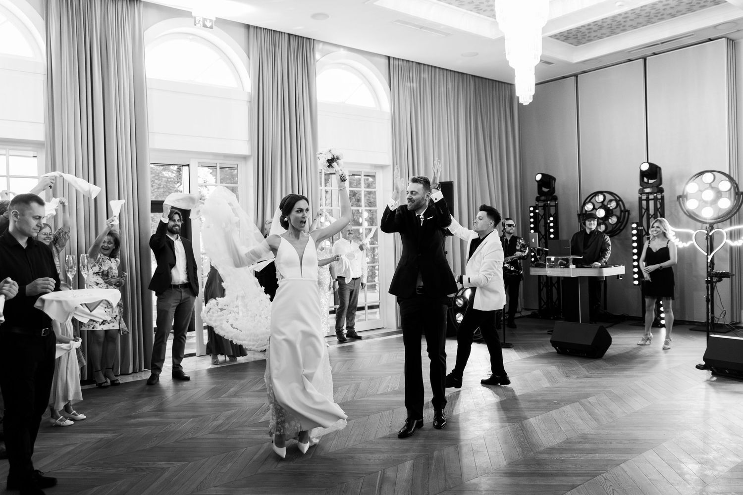 Wedding guests dancing and celebrating in an elegant ballroom with high ceilings and large windows.