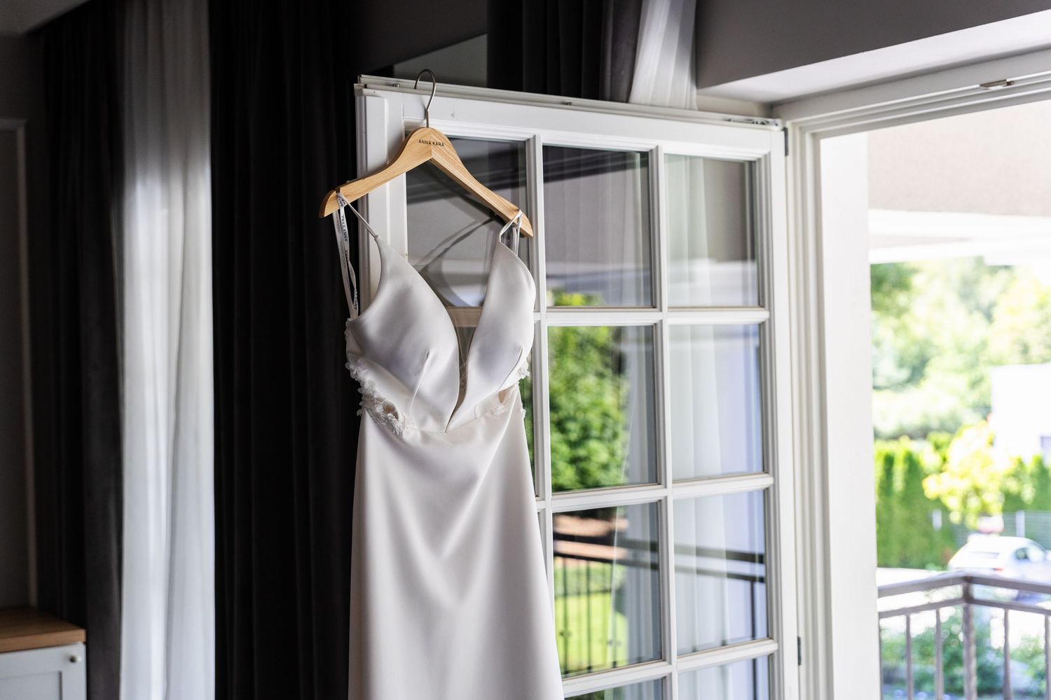 Elegant silver wedding dress hangs near bright window with wooden hanger in natural light.