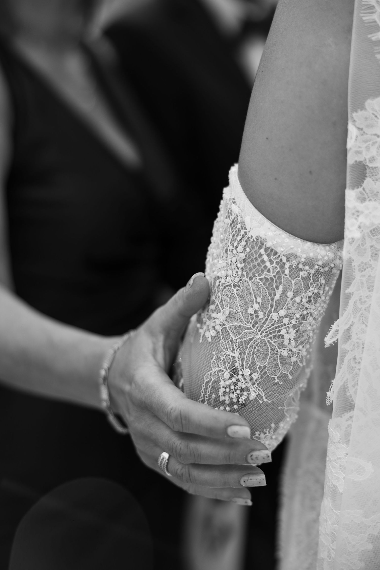 Black and white photo of hands adjusting a bride's lace wedding dress sleeve with delicate floral embroidery.