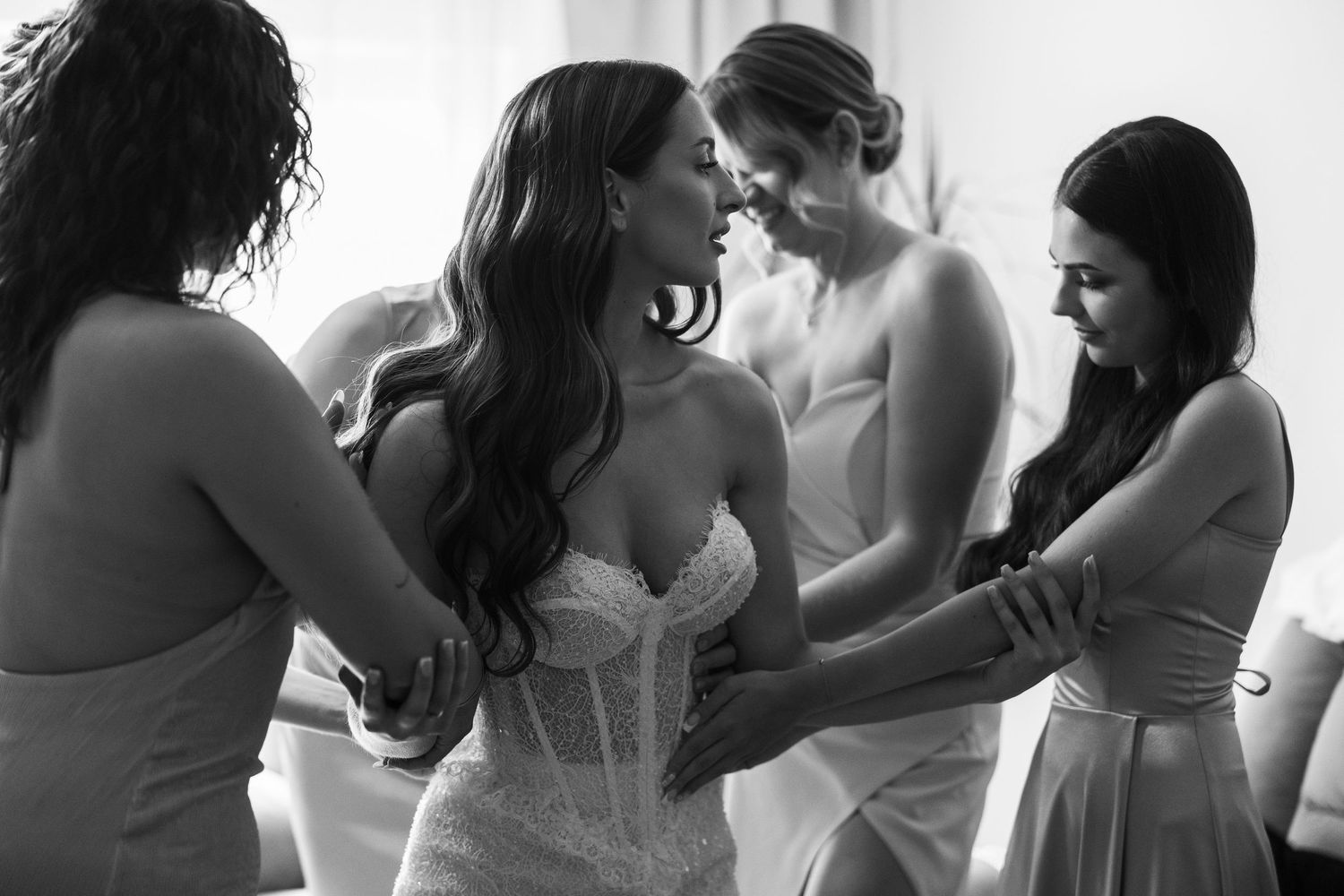 Black and white photo of bride in lace corset gown being helped by bridesmaids in elegant dresses during wedding preparation.