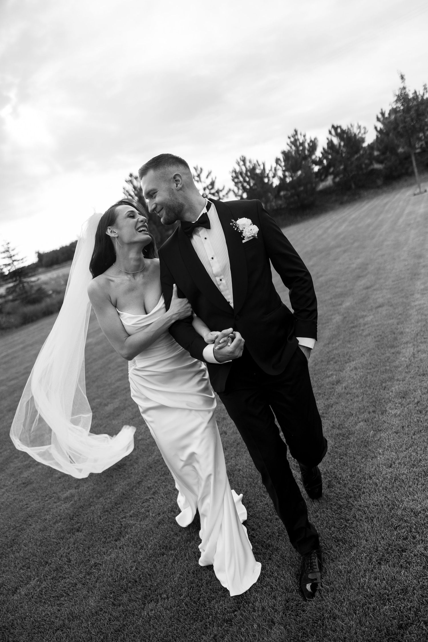 Black and white photo of a bride and groom walking together outdoors, smiling, bride in white gown with veil, groom in tuxedo.