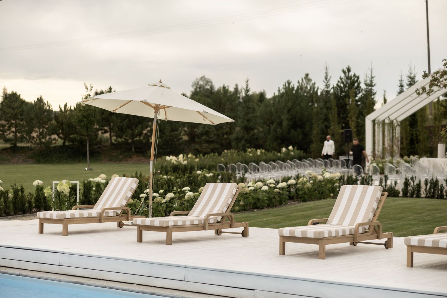Elegant poolside lounge chairs with striped cushions and a white umbrella beside a lush garden landscape.