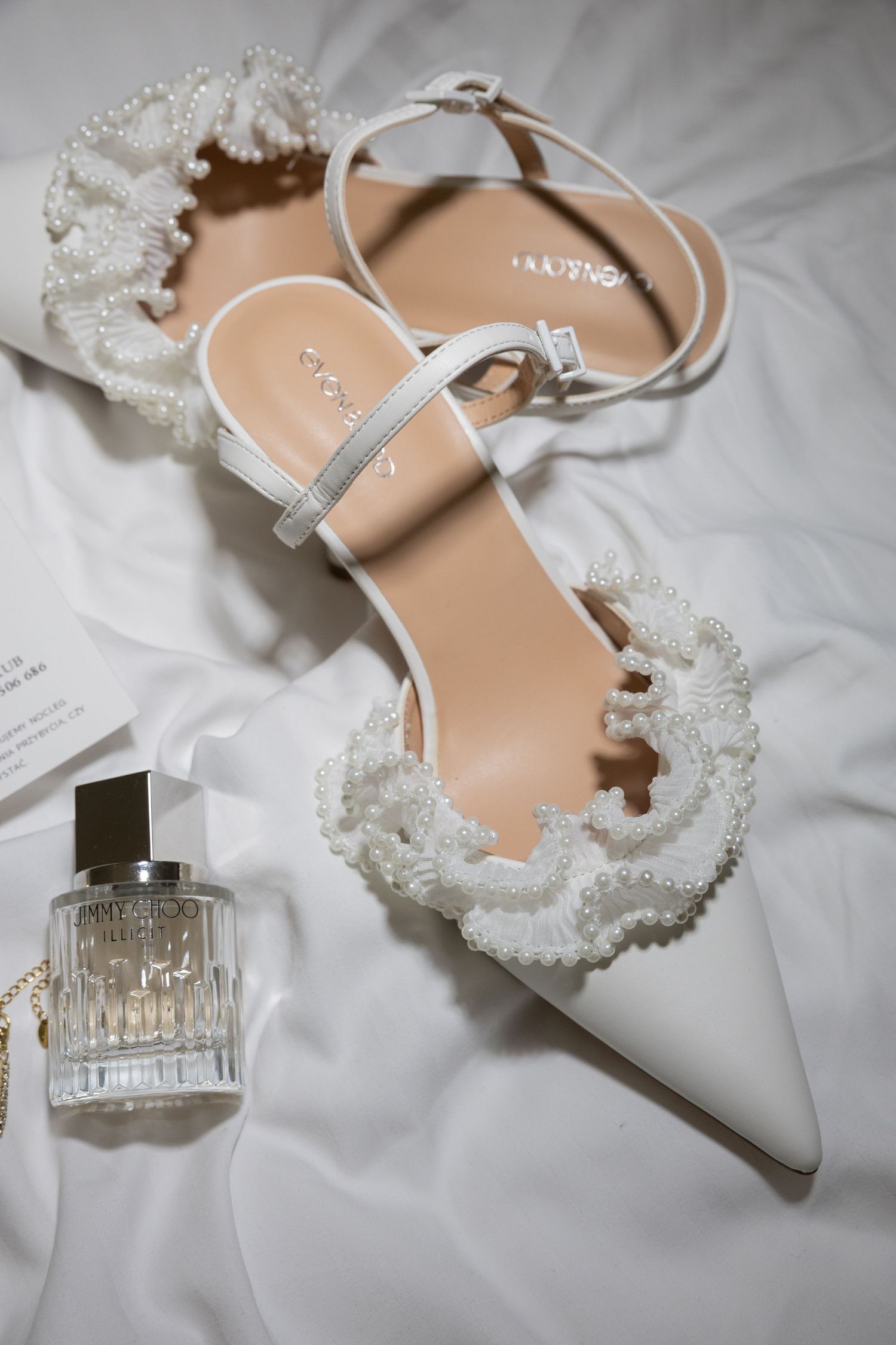 White bridal heels with pearl lace detail and ankle strap beside a perfume bottle on white fabric.