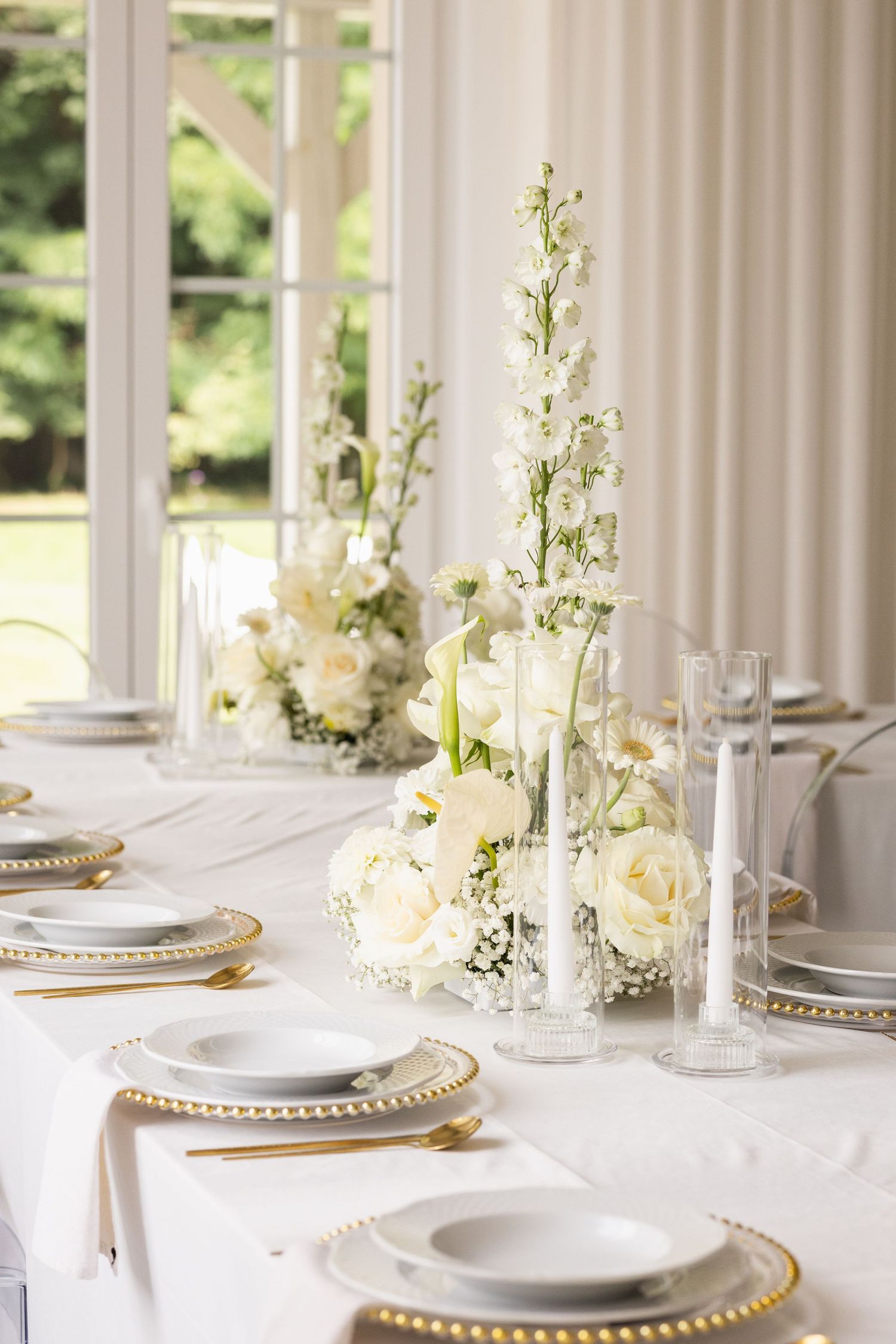 Elegant white table setting with gold charger plates, white flowers in vases, and natural light streaming through windows.