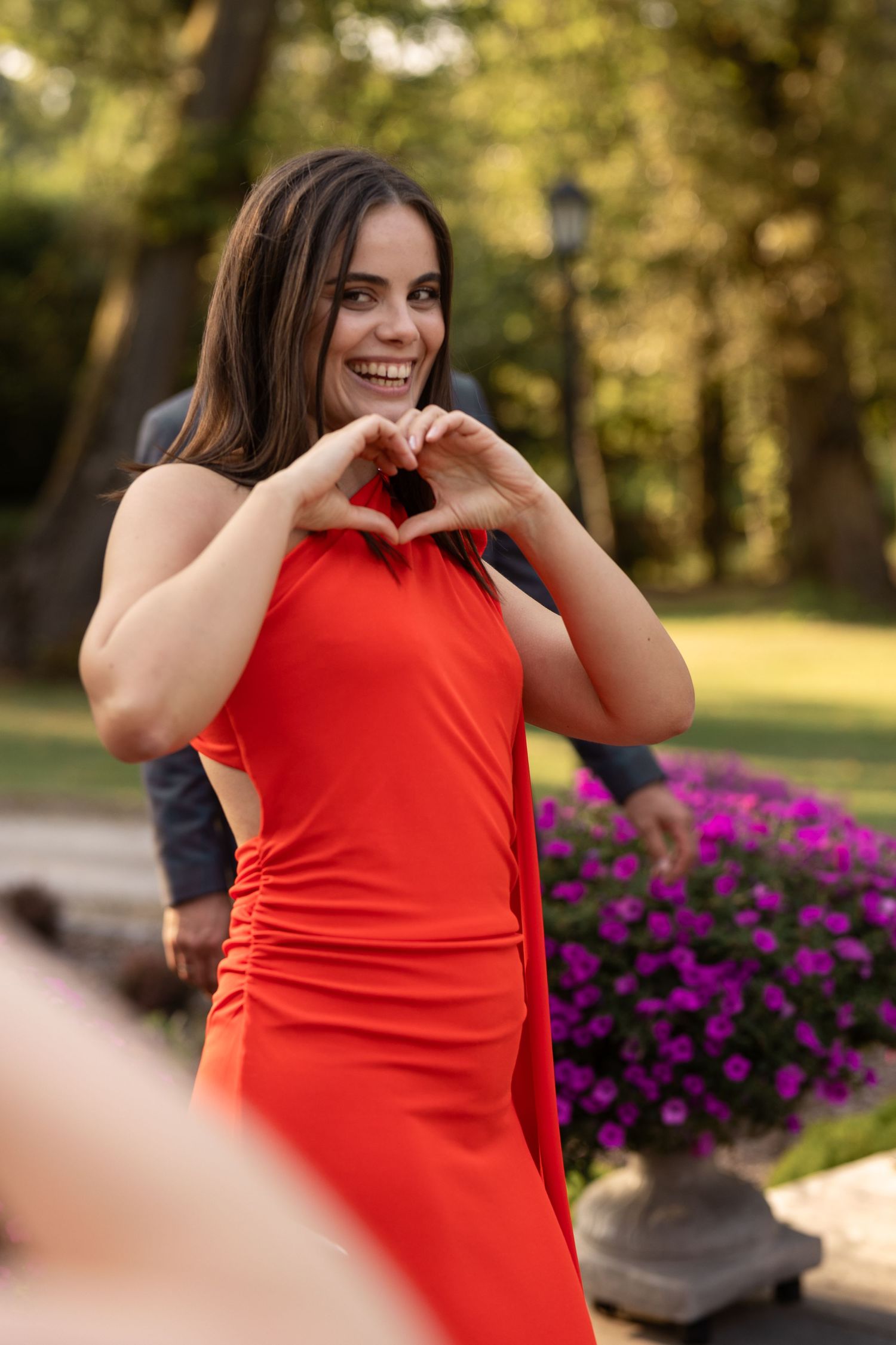 Woman in bright red dress poses playfully in a garden setting with purple flowers in the background.