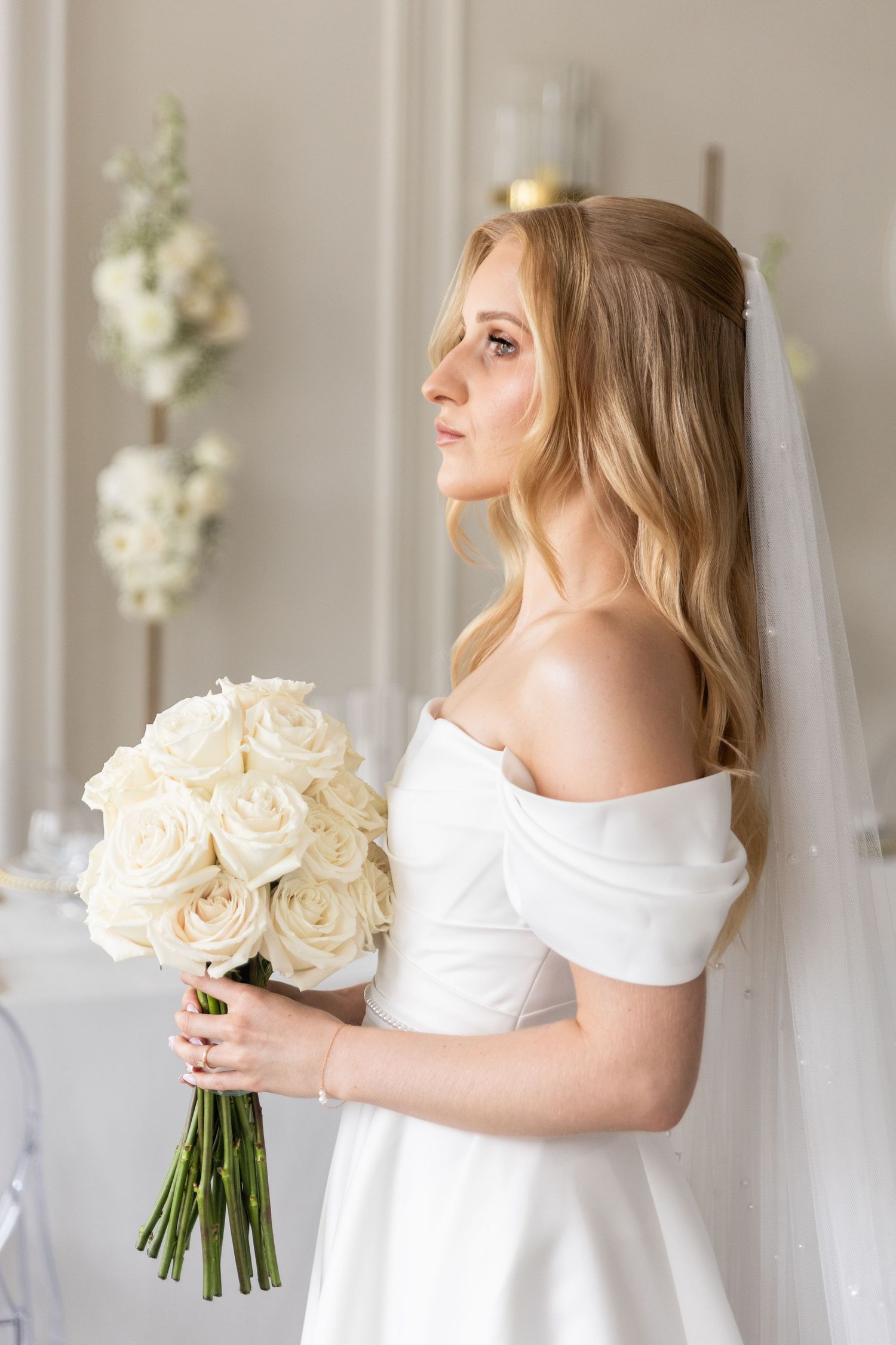 A bride in a white off-shoulder dress holds an elegant bouquet of cream roses in a light-filled room.