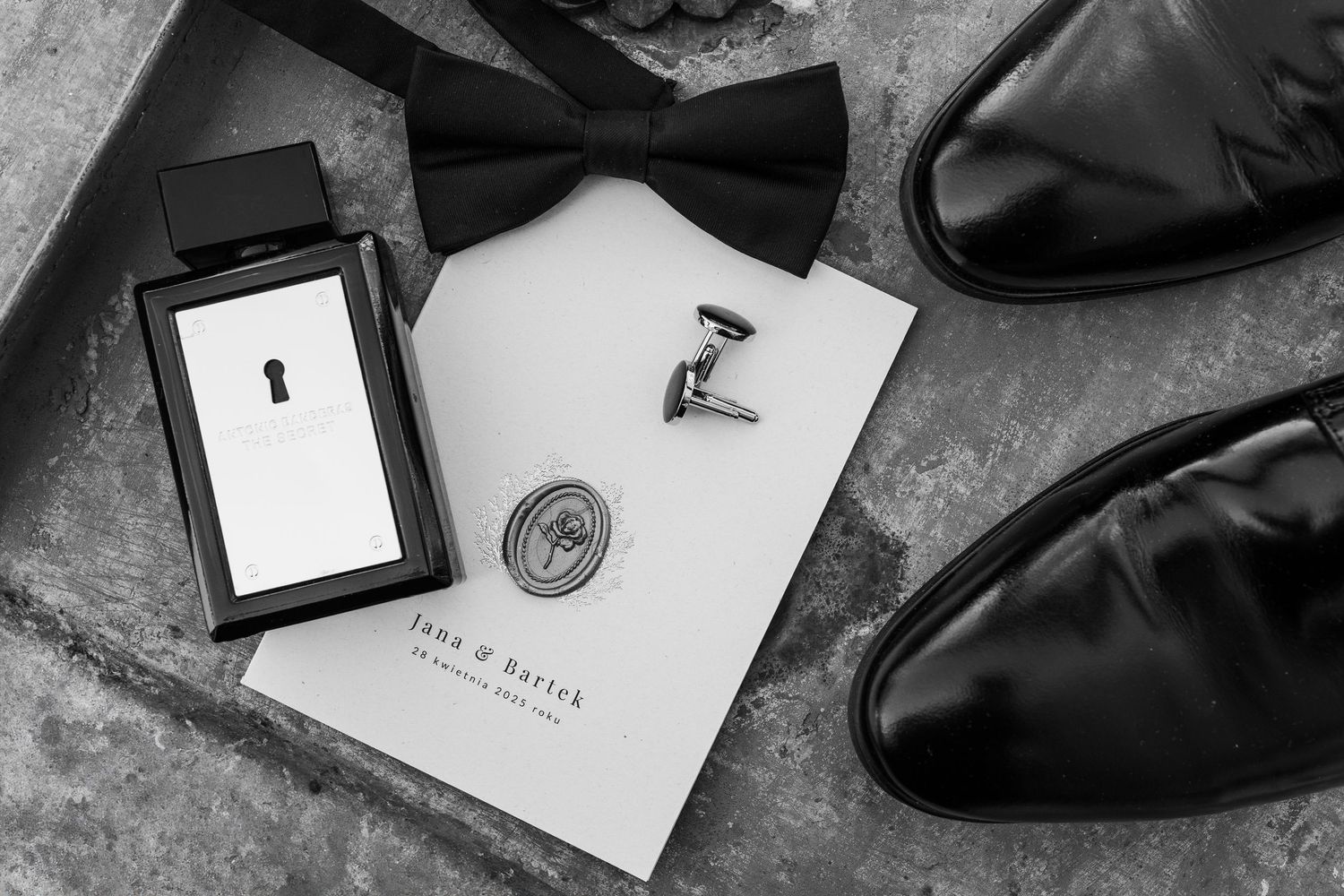 Black and white photo of groom's accessories including bow tie, dress shoes, cufflinks and small frame laid on concrete surface.