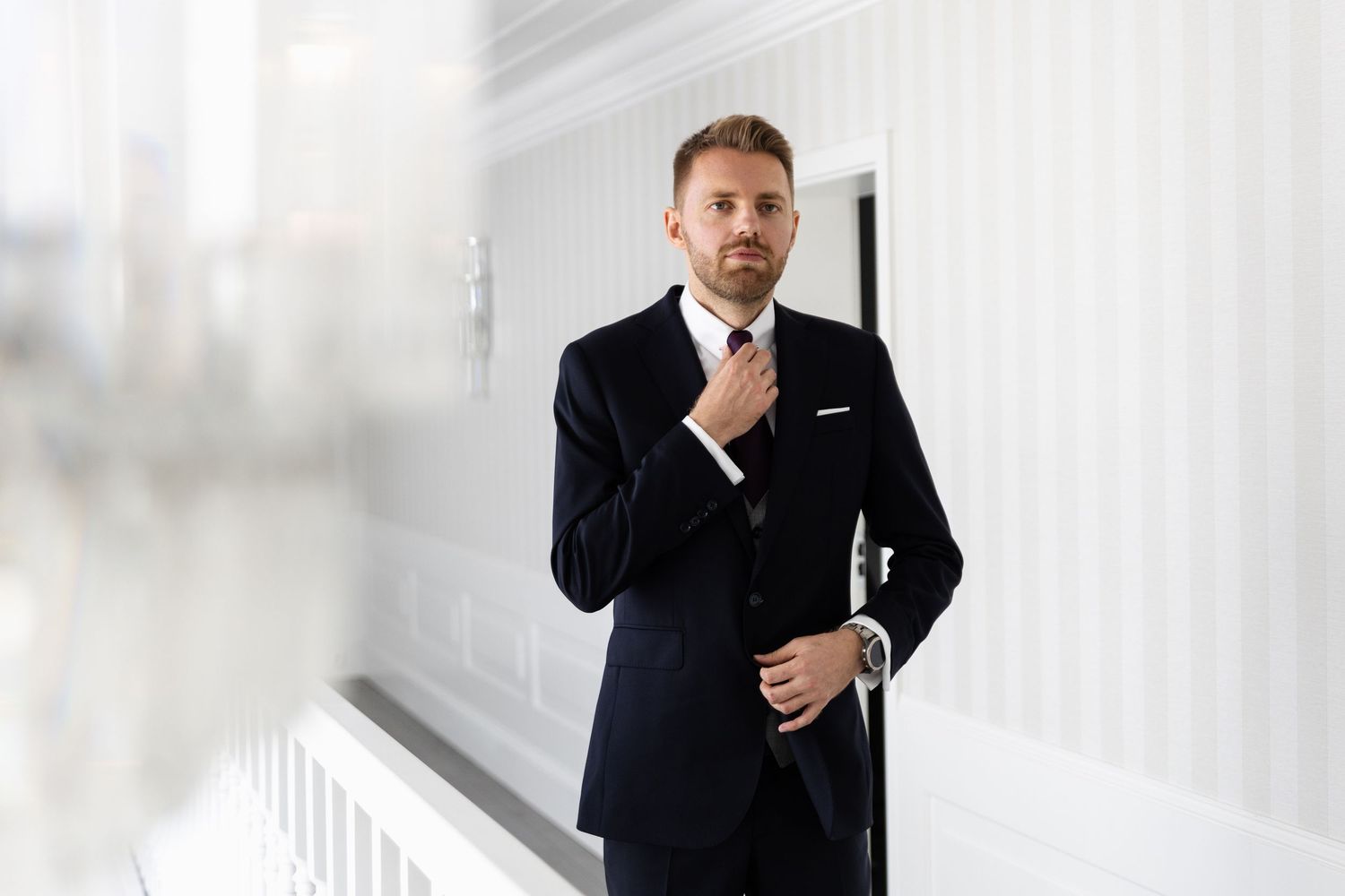 Man in black suit adjusts his tie while standing in elegant white interior hallway.