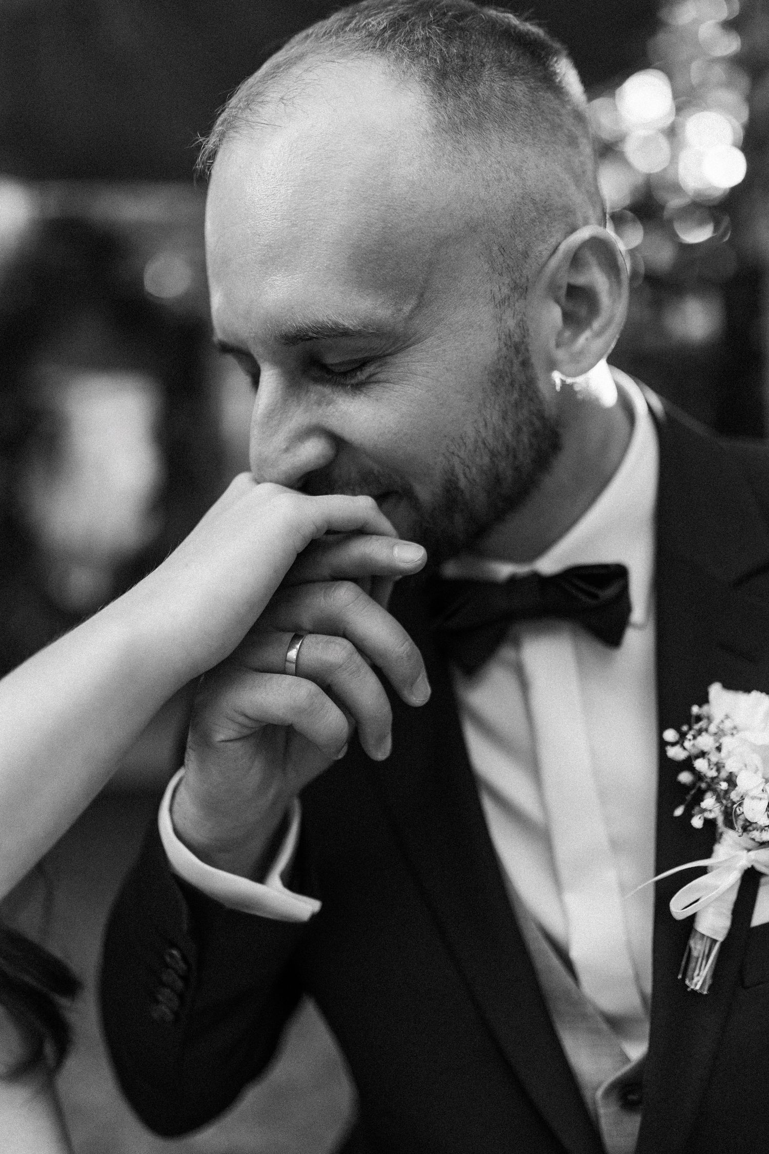 Black and white candid wedding photo showing emotional moment with groom kissing hand while wearing tuxedo and boutonniere.