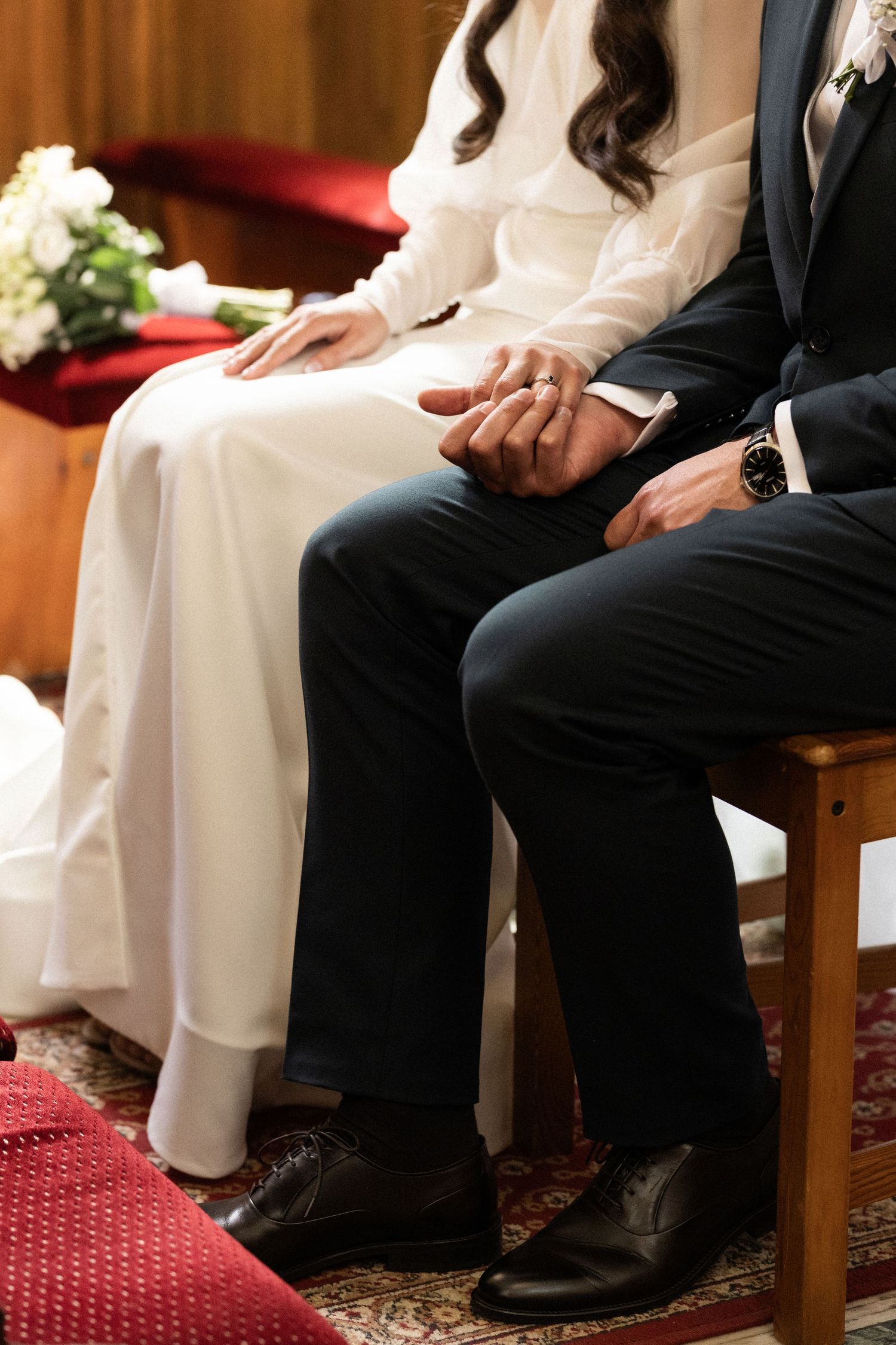 A couple holds hands while sitting together during their wedding ceremony on wooden chairs with a red carpet beneath them.