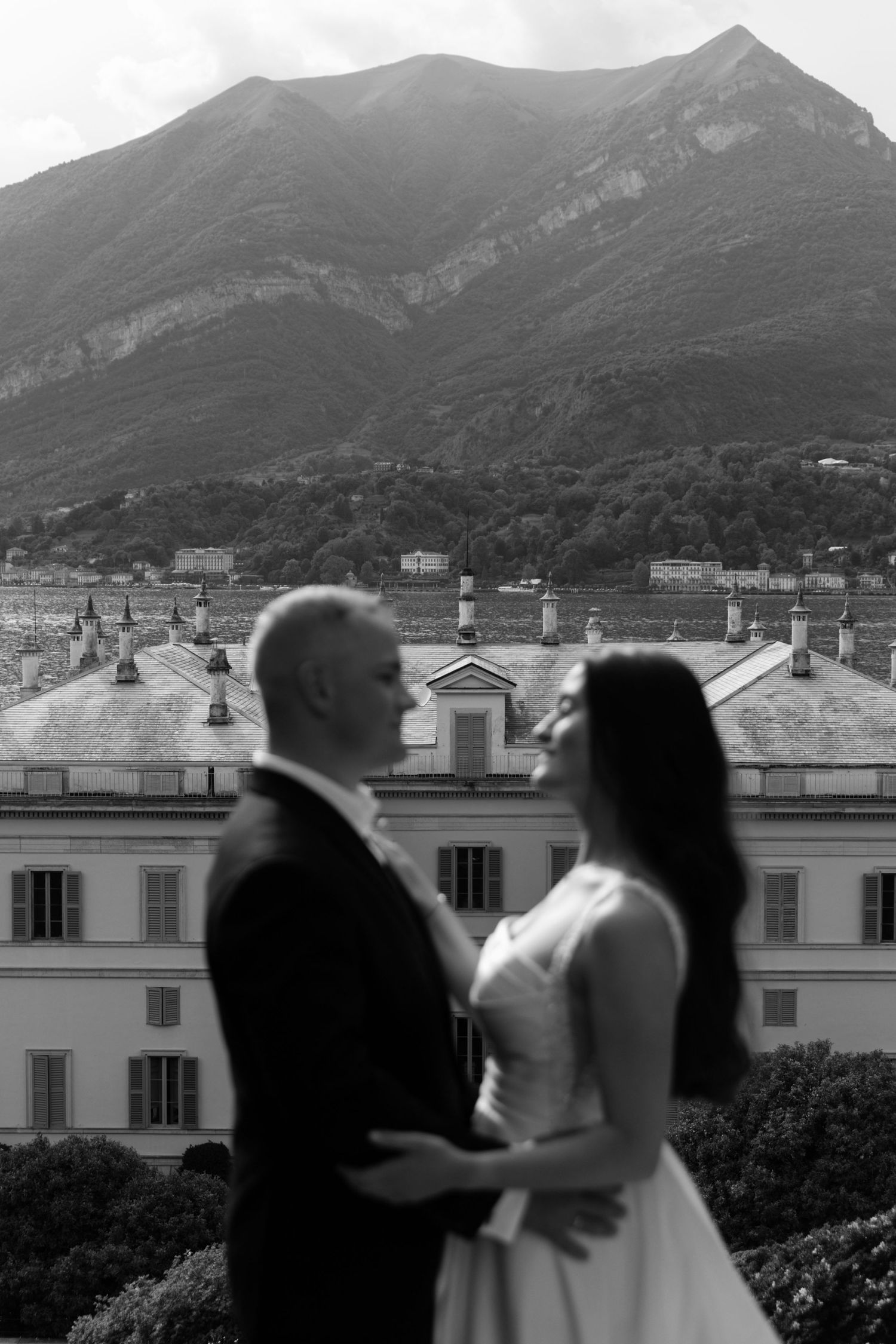 Black and white wedding photo of couple embracing with Italian villa and mountain backdrop at Lake Como.