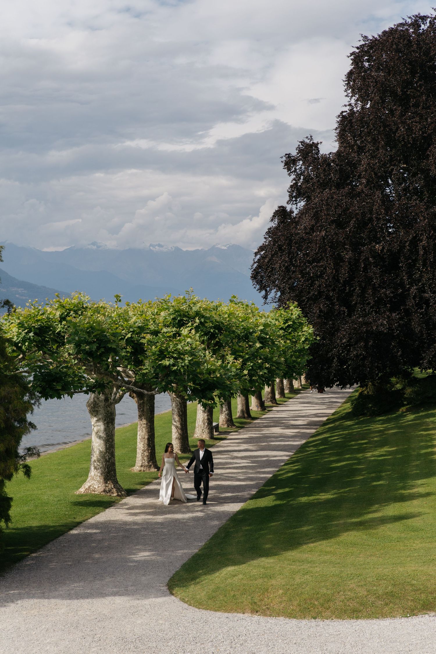 Bride and groom walking along a tree-lined path at an elegant lakeside estate with mountain views.
