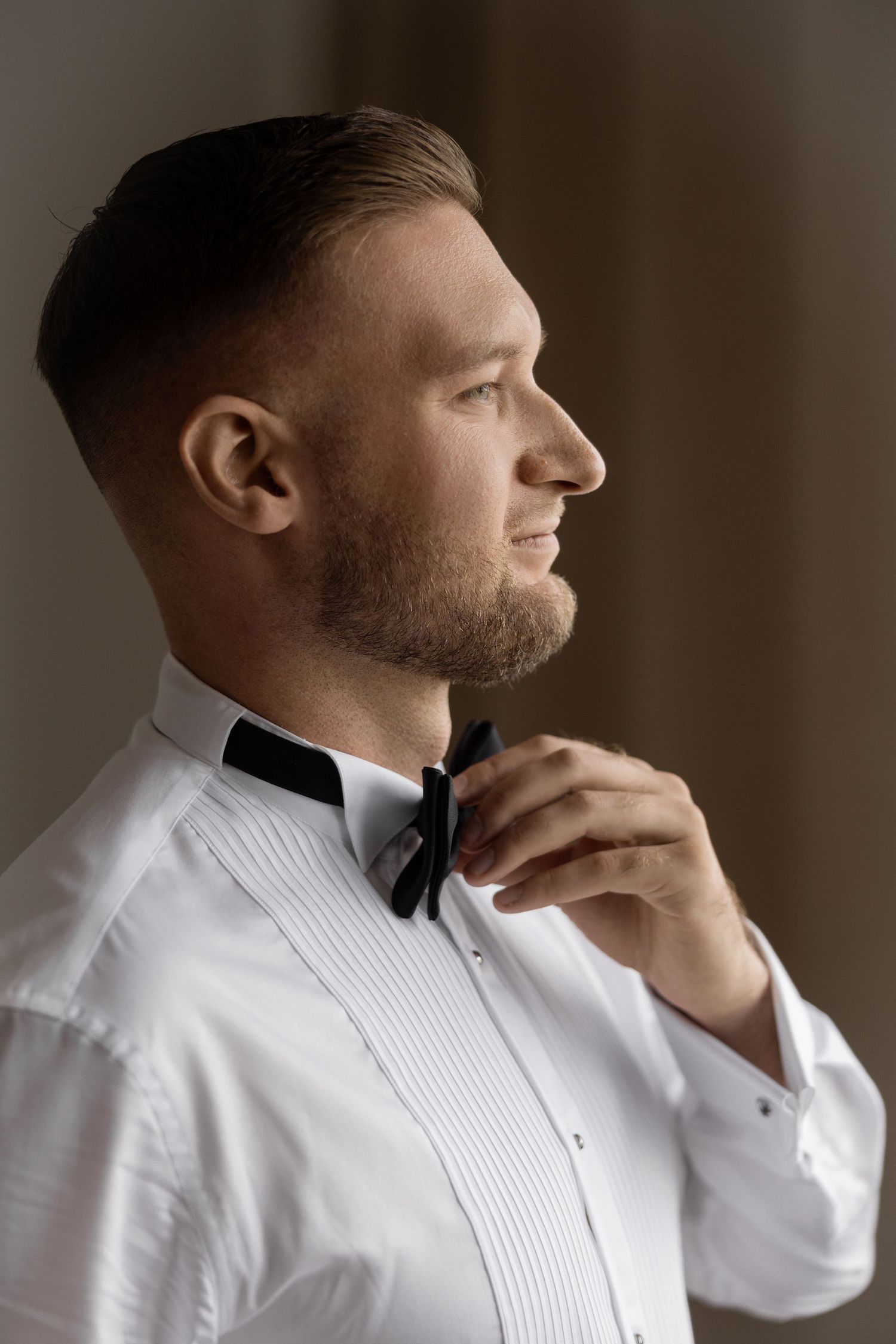 A well-dressed man in a white tuxedo shirt adjusting his black bow tie, captured in a side profile portrait.