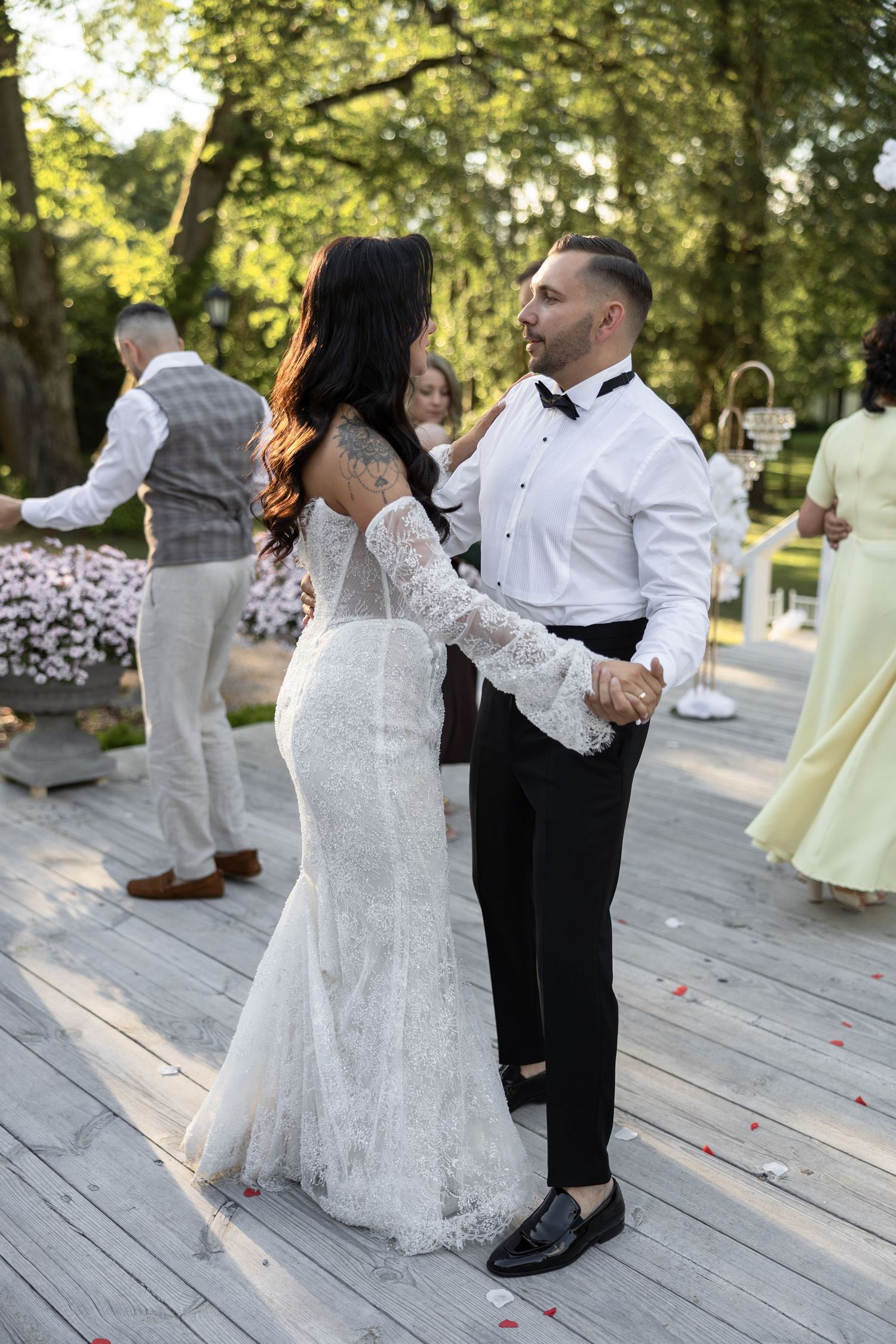 Bride and groom share their first dance outdoors on a wooden deck surrounded by guests and lush green trees.