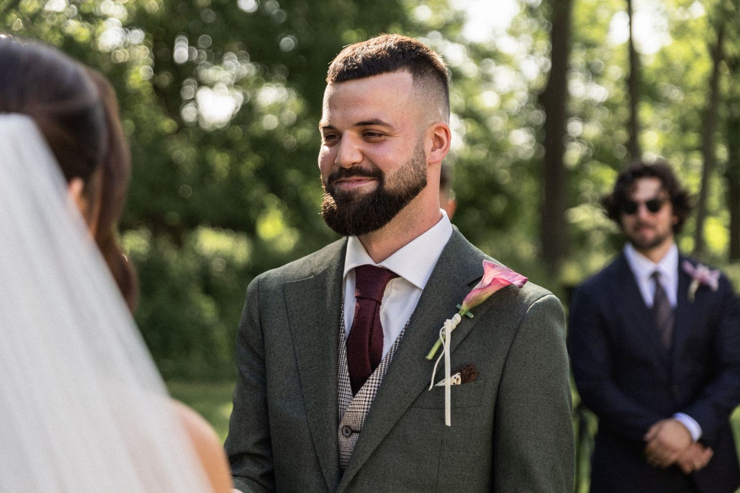 Groom wearing a green suit with burgundy tie watches bride during outdoor wedding ceremony in sunlit forest setting.
