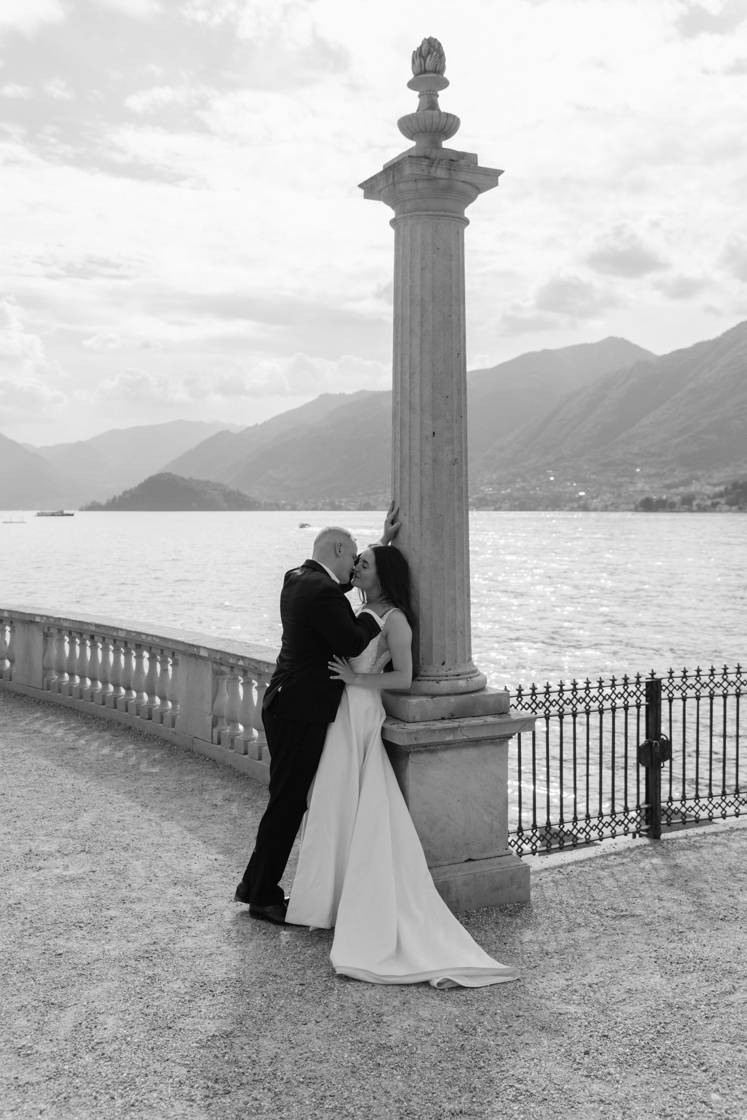 Black and white photo of a bride and groom kissing by a stone column on a lakeside terrace with mountains in the background.