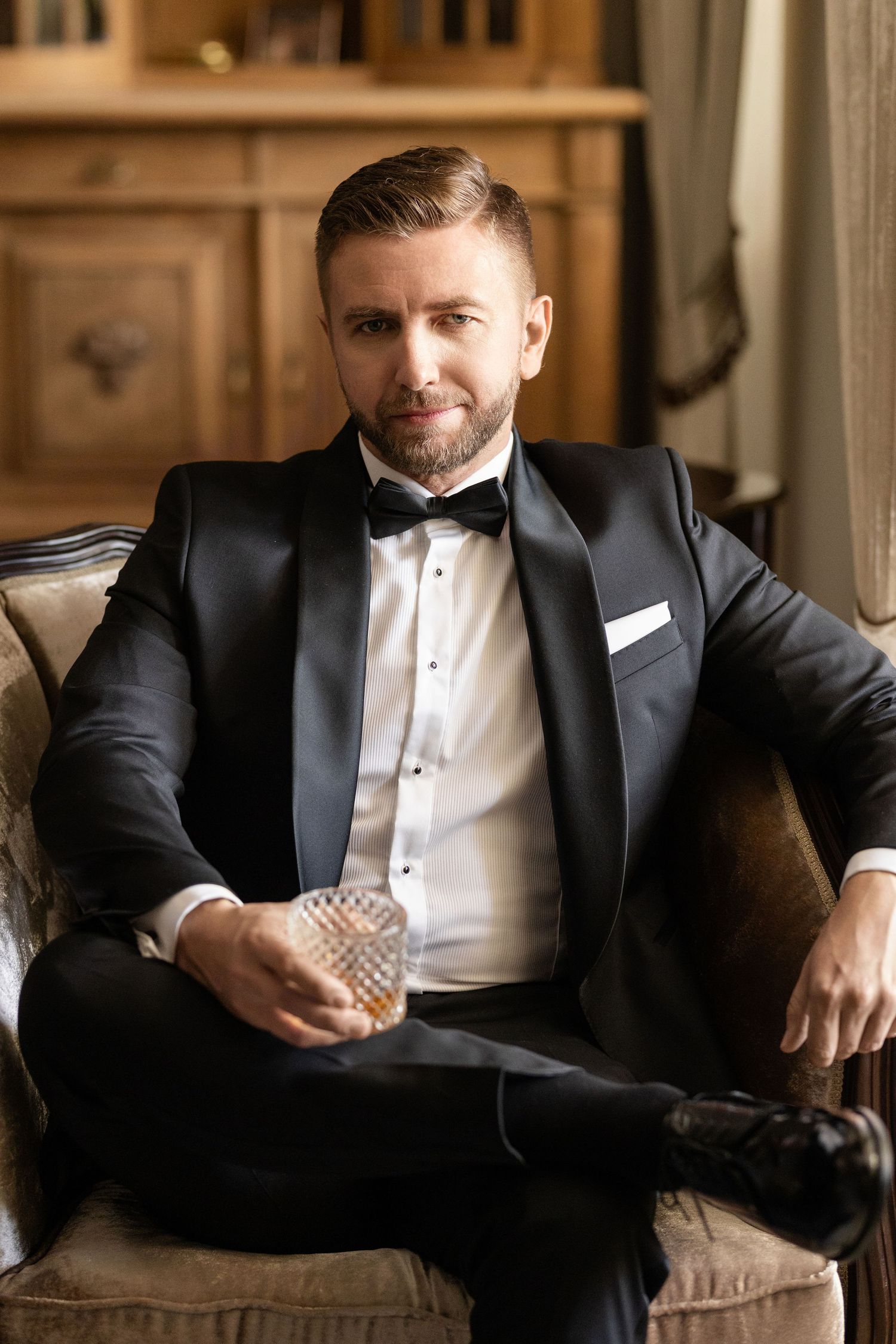 Elegant groom in black tuxedo and white bow tie sits on vintage brown leather chair in classic setting.