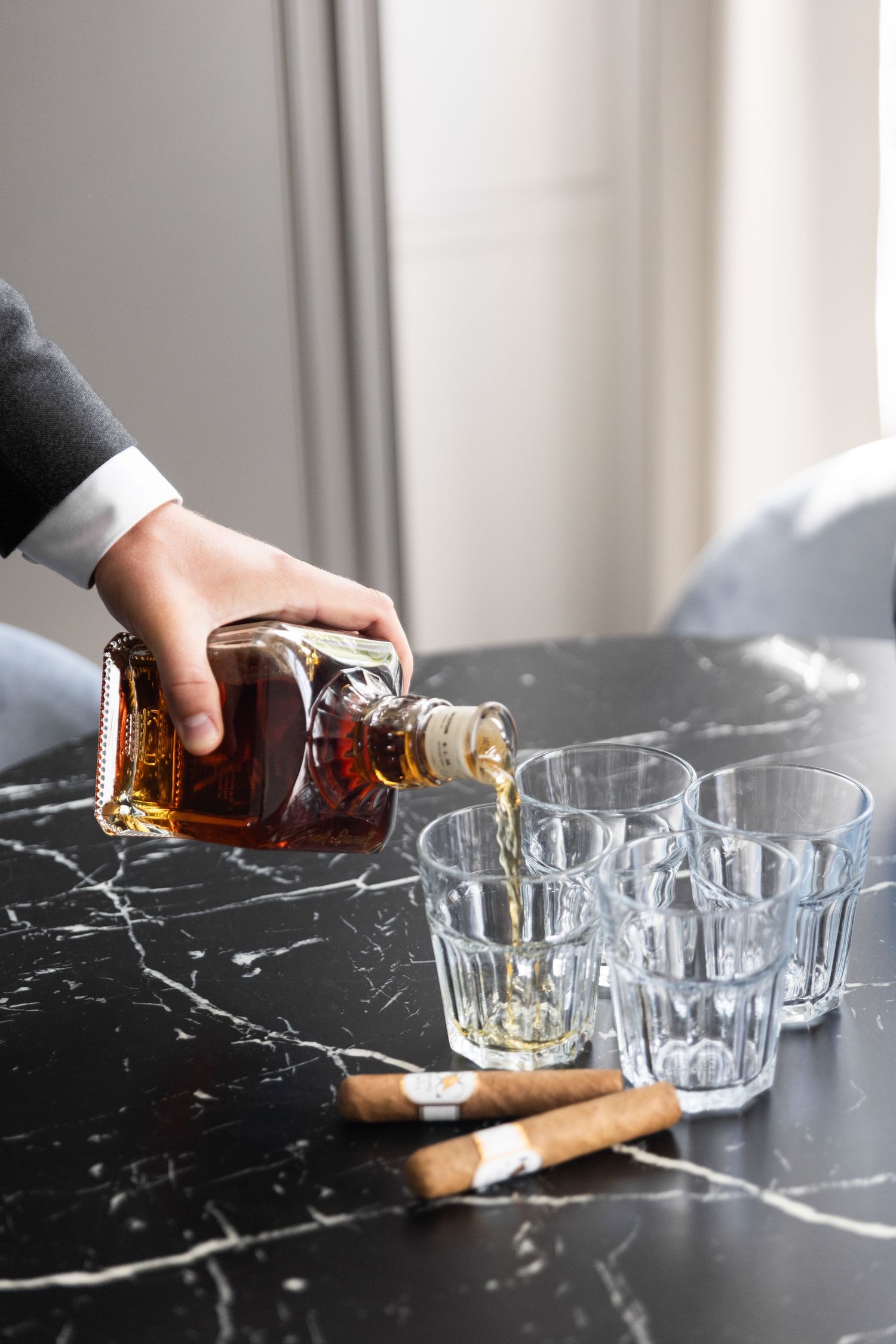 Hands pouring whiskey into crystal glasses on a black marble surface with a cigar in the scene.
