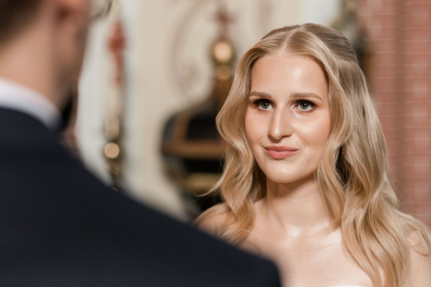 Bride with wavy blonde hair smiles lovingly during wedding ceremony with soft lighting and brick wall backdrop.