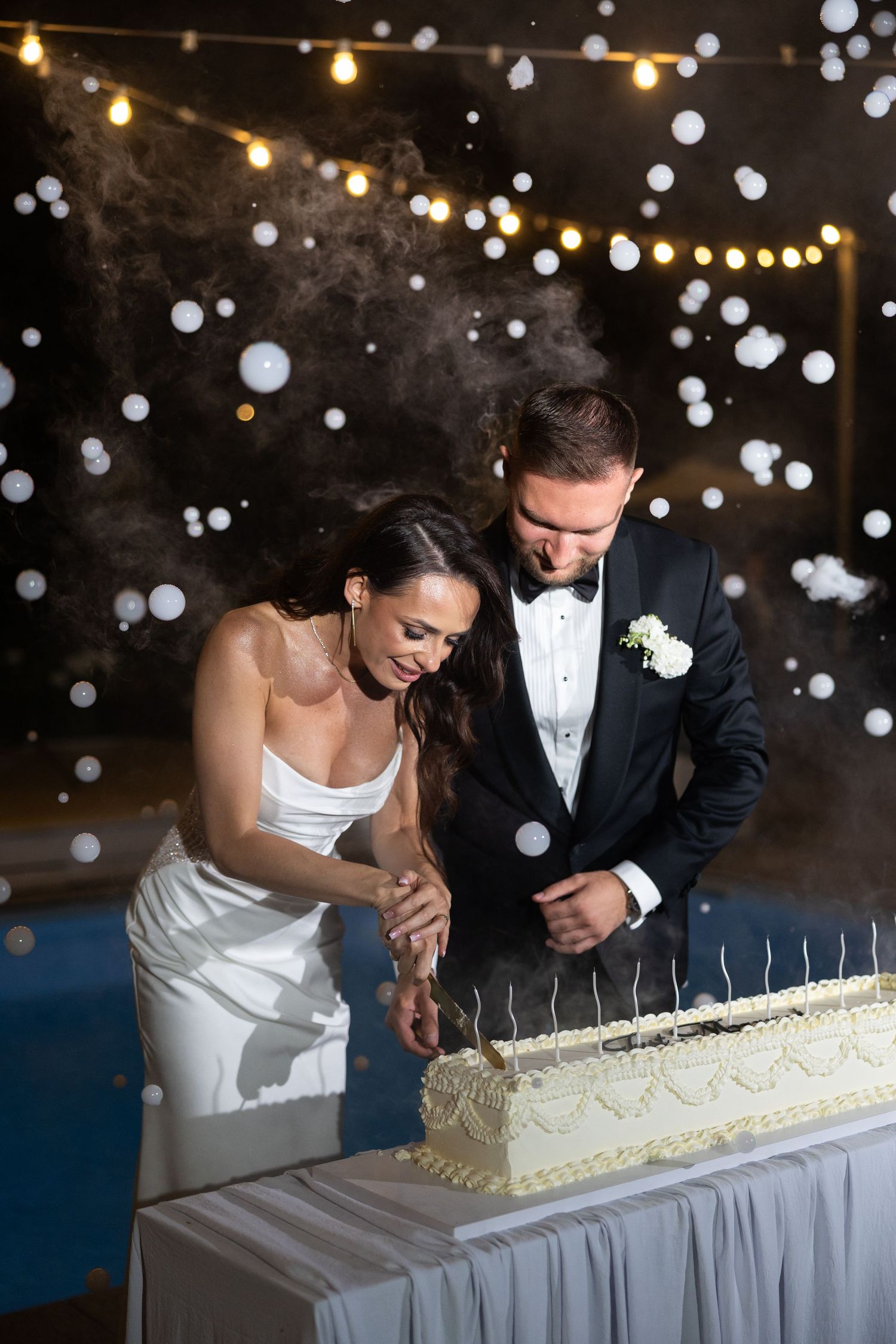 Bride and groom cutting their wedding cake together at a romantic evening reception with bokeh lights in the background.