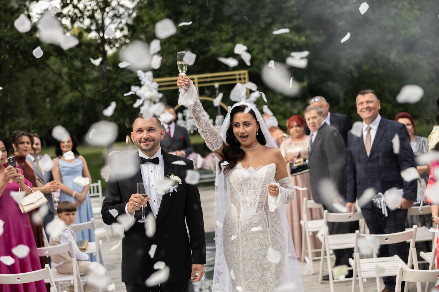 Joyful newlyweds walk down the aisle as white petals fill the air at an outdoor wedding ceremony.