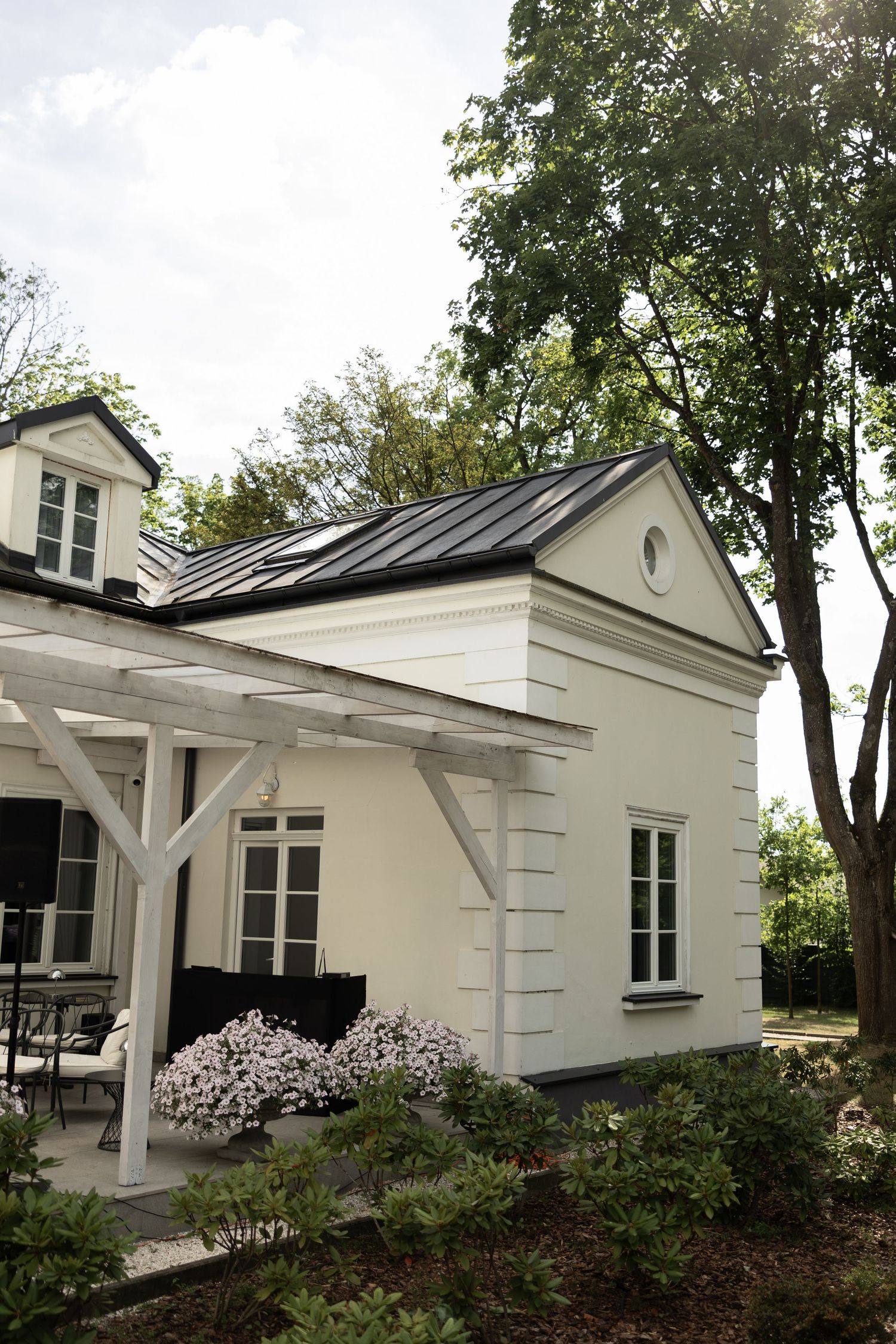 White classic house with dark metal roof, pergola, and blooming garden surrounded by tall trees on a sunny day.