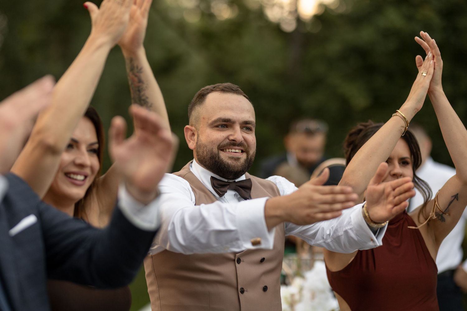 Smiling groom in bow tie clapping with wedding guests outdoors at a festive celebration.