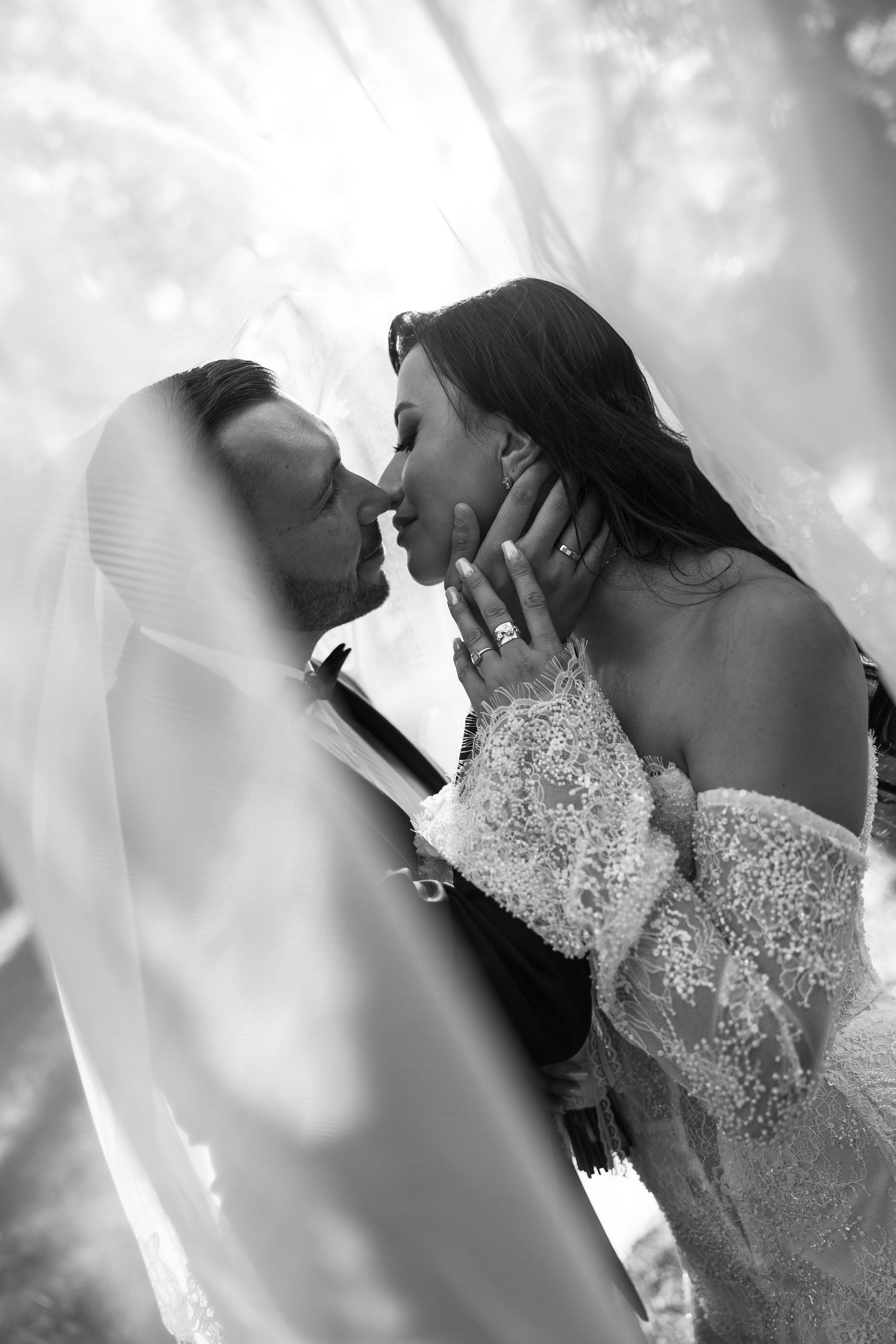 Black and white photo of bride and groom sharing a romantic kiss under a flowing veil on their wedding day.
