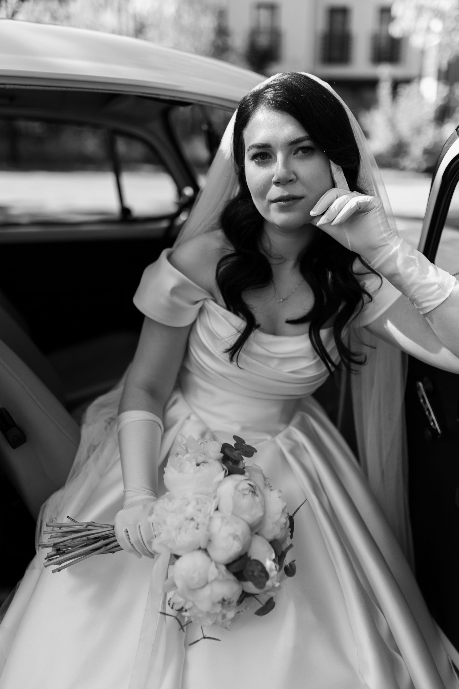 Black and white photo of a bride in an elegant white gown with long sleeves adjusting her veil while holding a peony bouquet.