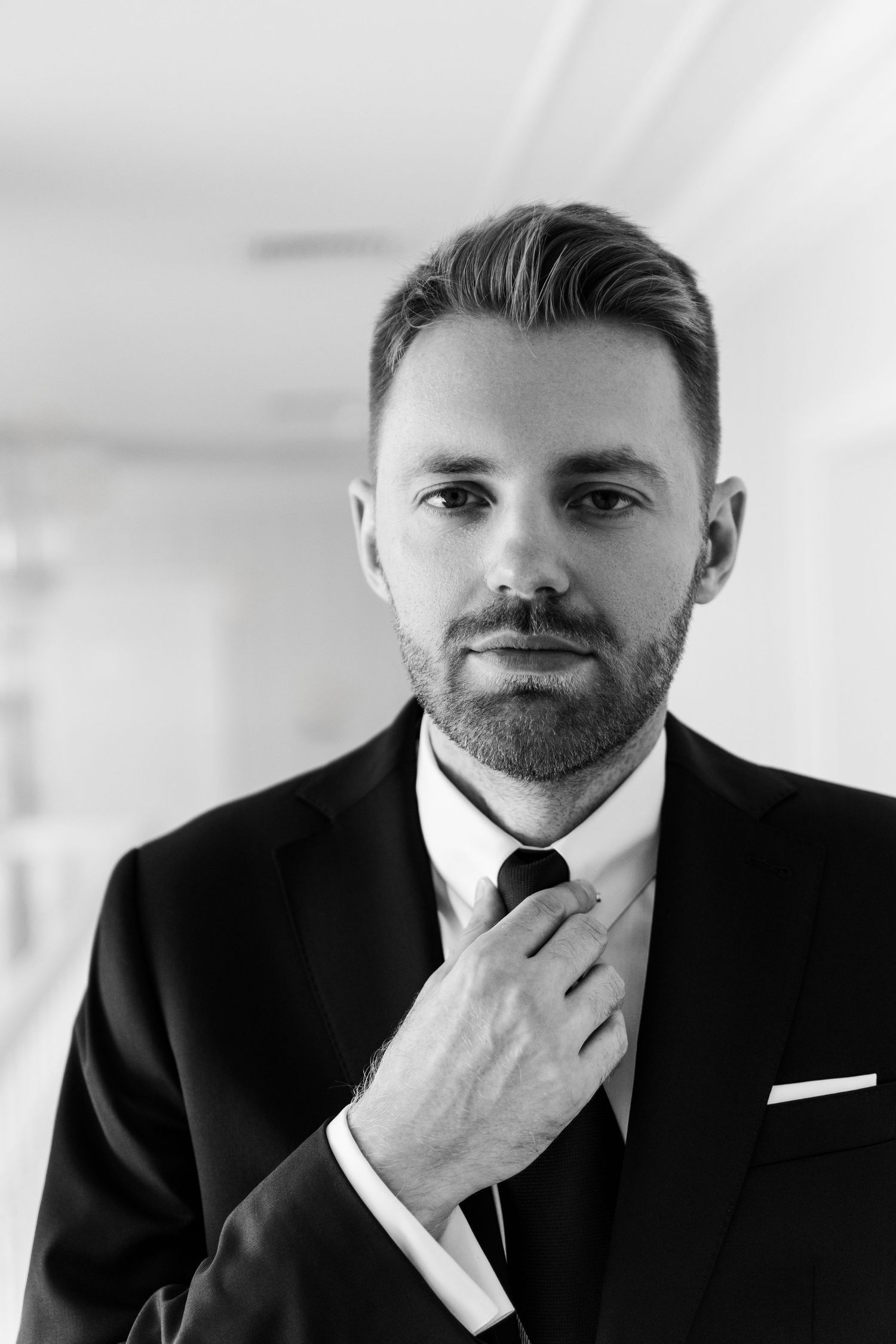 A black and white portrait of a well-groomed professional adjusting his tie while wearing a formal black suit.