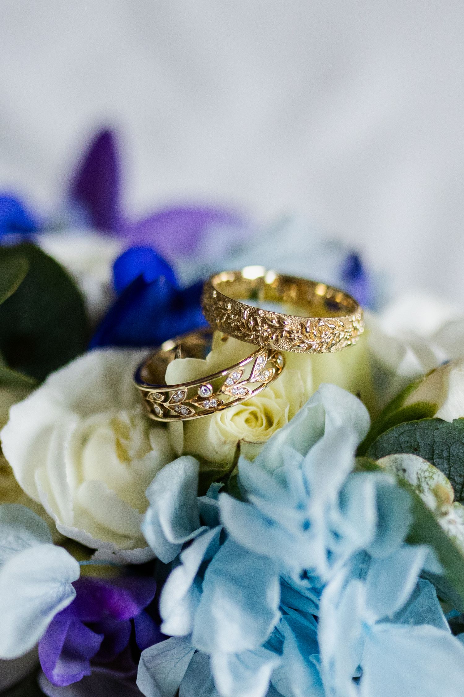 Two gold wedding rings with diamond accents rest on a bouquet of white, blue and purple flowers.