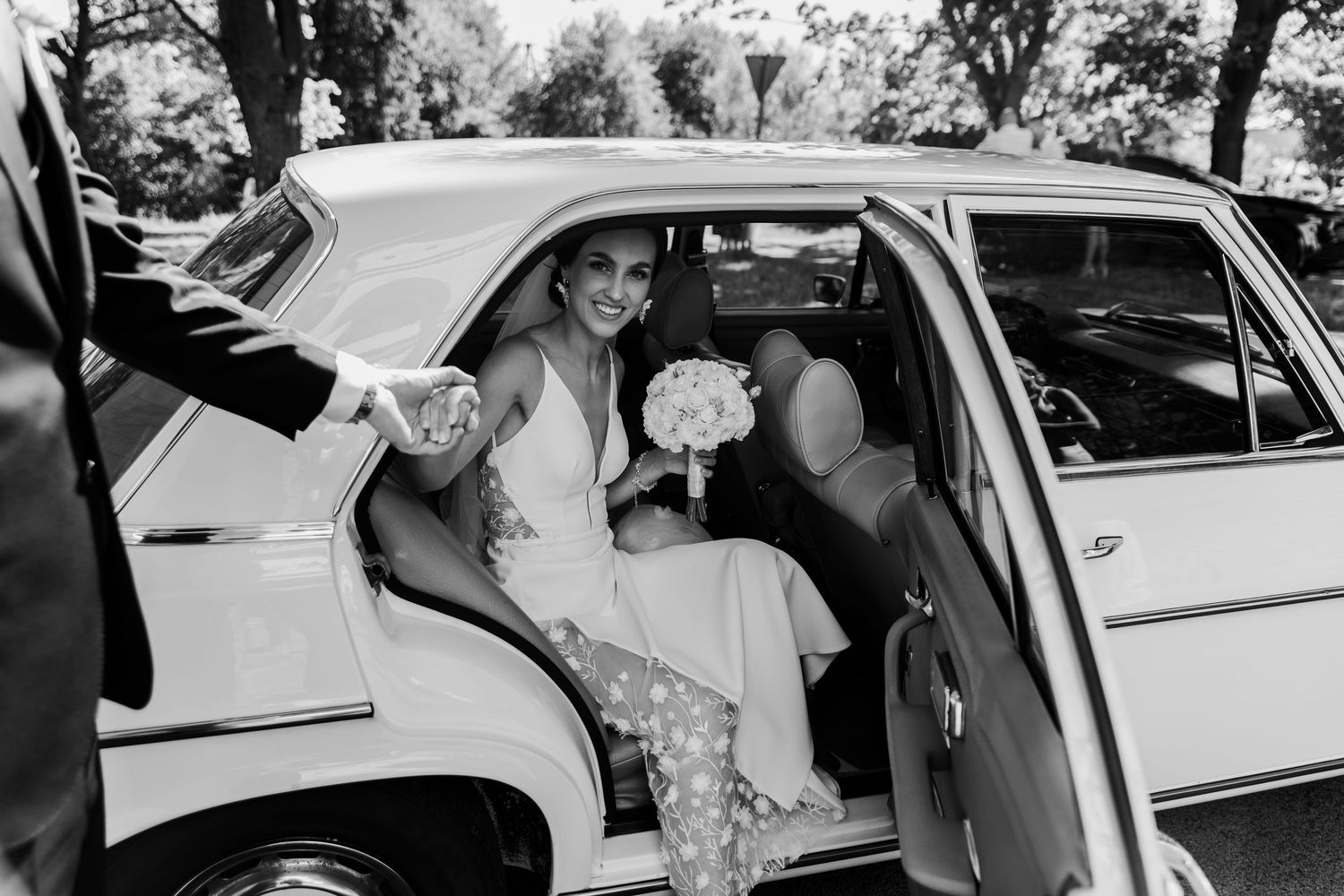 Classic vintage car with someone in a white gown and bouquet of flowers entering through the door in black and white.